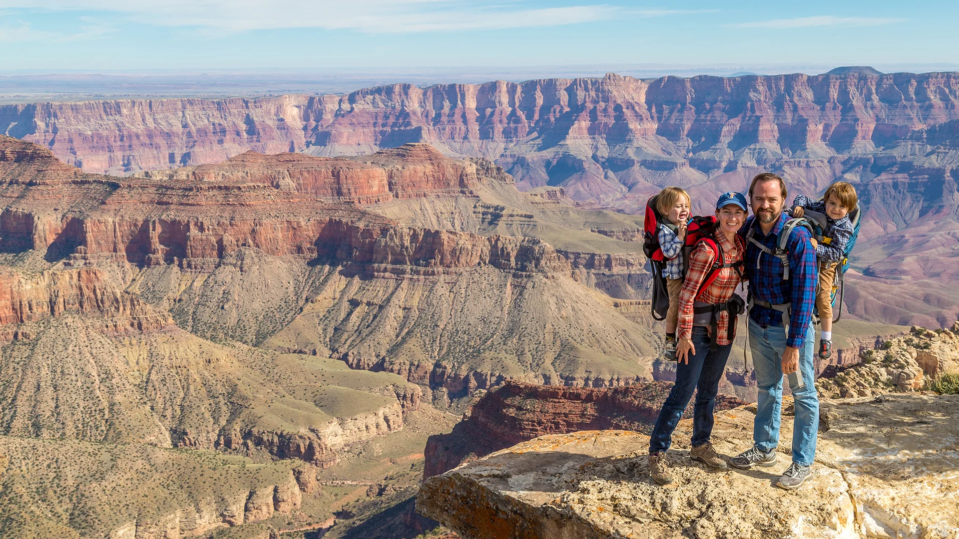 Family hiking at the Grand Canyon, parents smiling with young children in backpacks, overlooking vast canyon landscape under a clear sky.