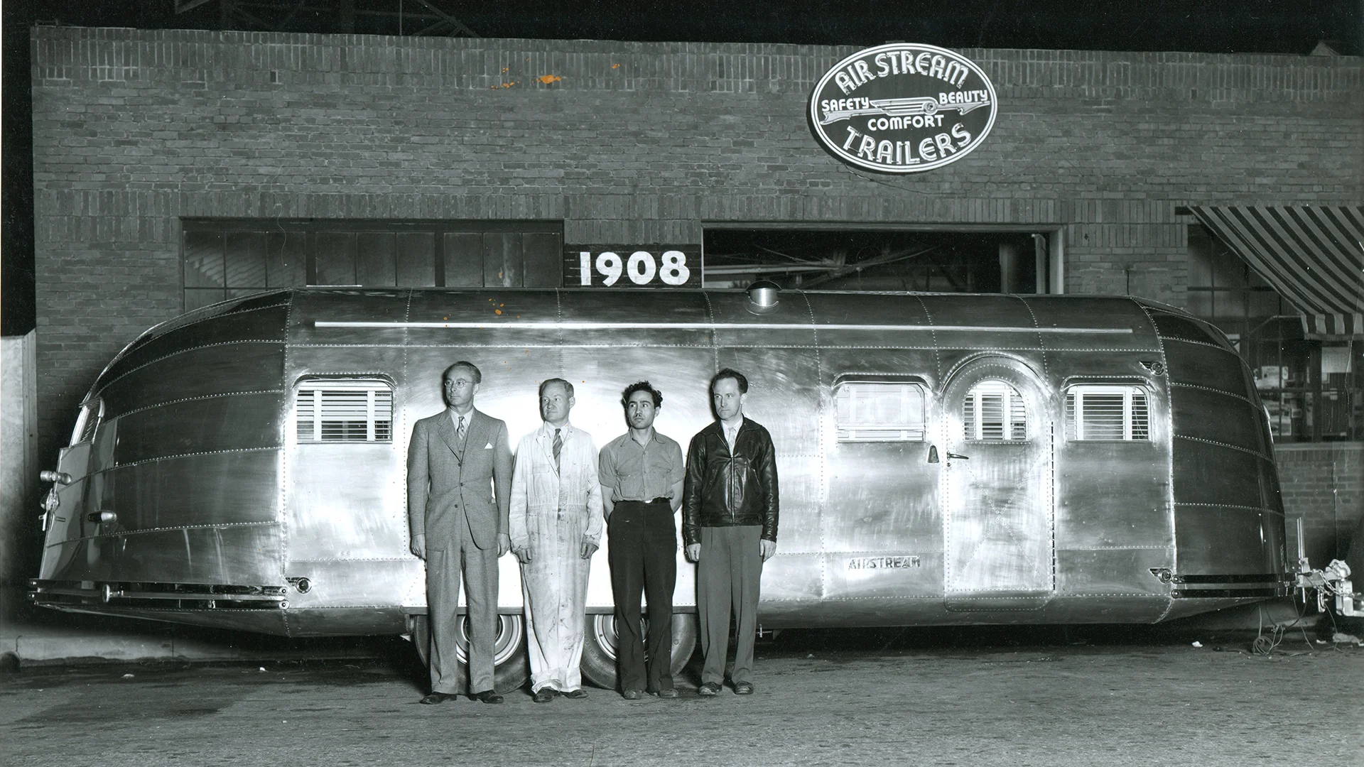 Four men stand in front of a shiny vintage Airstream trailer outside a brick building with a sign reading Airstream Trailers. The photo appears to be taken at night.