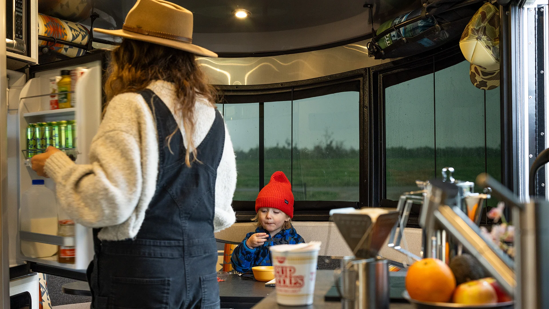 Woman in a hat opens a fridge in an RV kitchen, while a child in a red beanie eats at the table. Large windows show a rainy landscape outside.