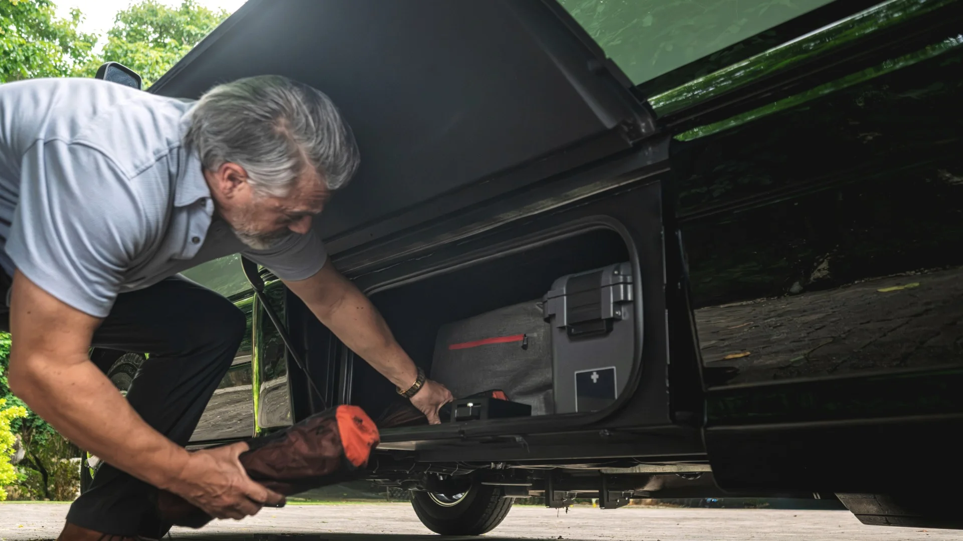 A man in a light shirt places an item into a storage compartment on the side of a black vehicle parked on a paved area with trees in the background.