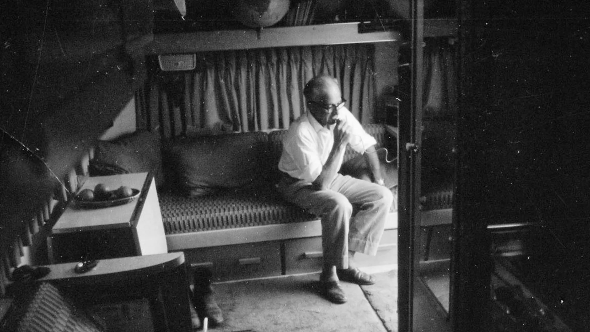 A person sits thoughtfully on a sofa in a dimly lit room, with a curtain and a globe visible in the background.