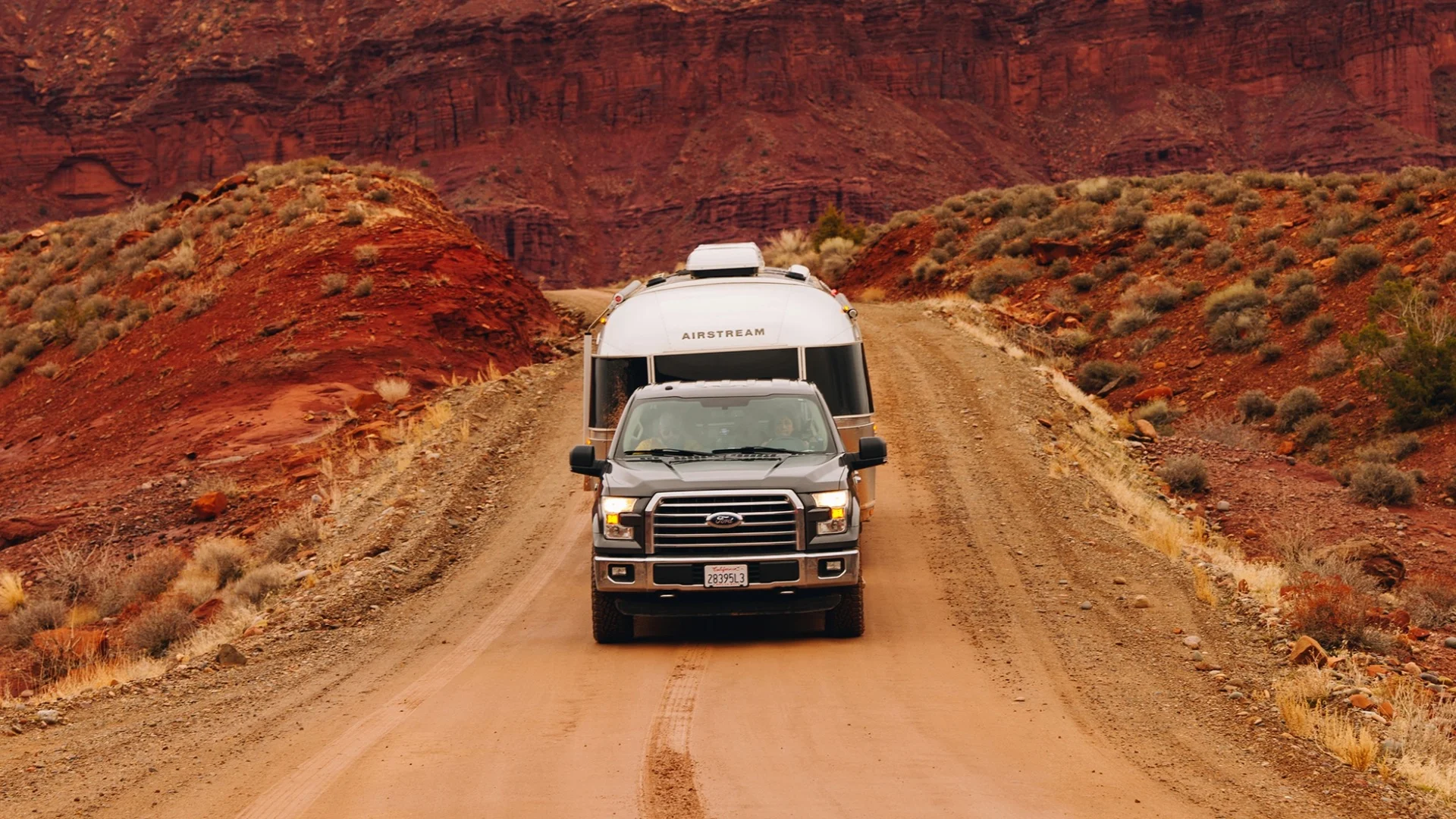A truck towing an Airstream trailer navigates a dirt road through a scenic, rugged red rock canyon landscape.