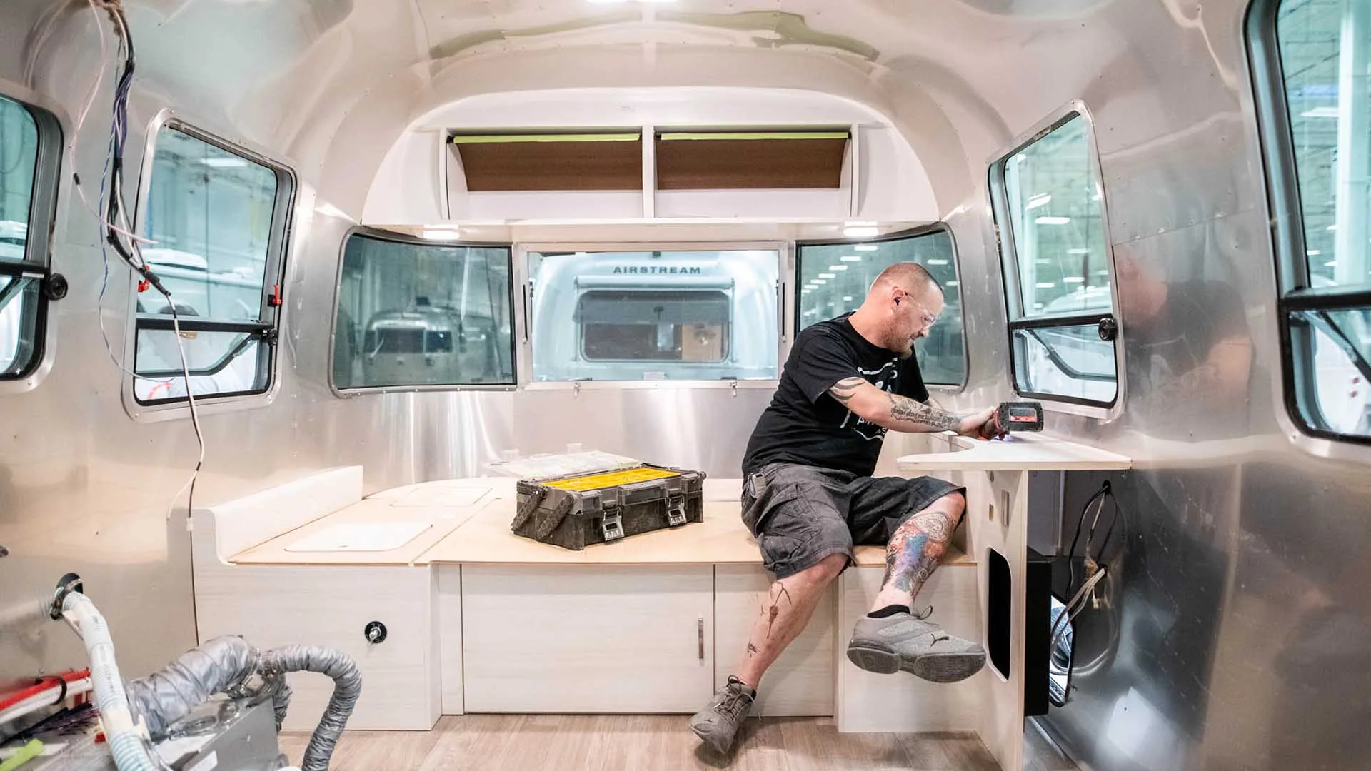 Man works in an Airstream interior, sitting on a built-in seat, using tools. Wires and boxes are visible, suggesting ongoing renovation or setup.