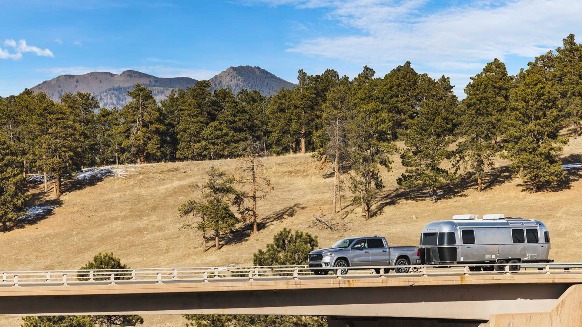 A silver truck towing an Airstream trailer crosses a bridge, with pine trees and a mountain range in the background under a clear blue sky.