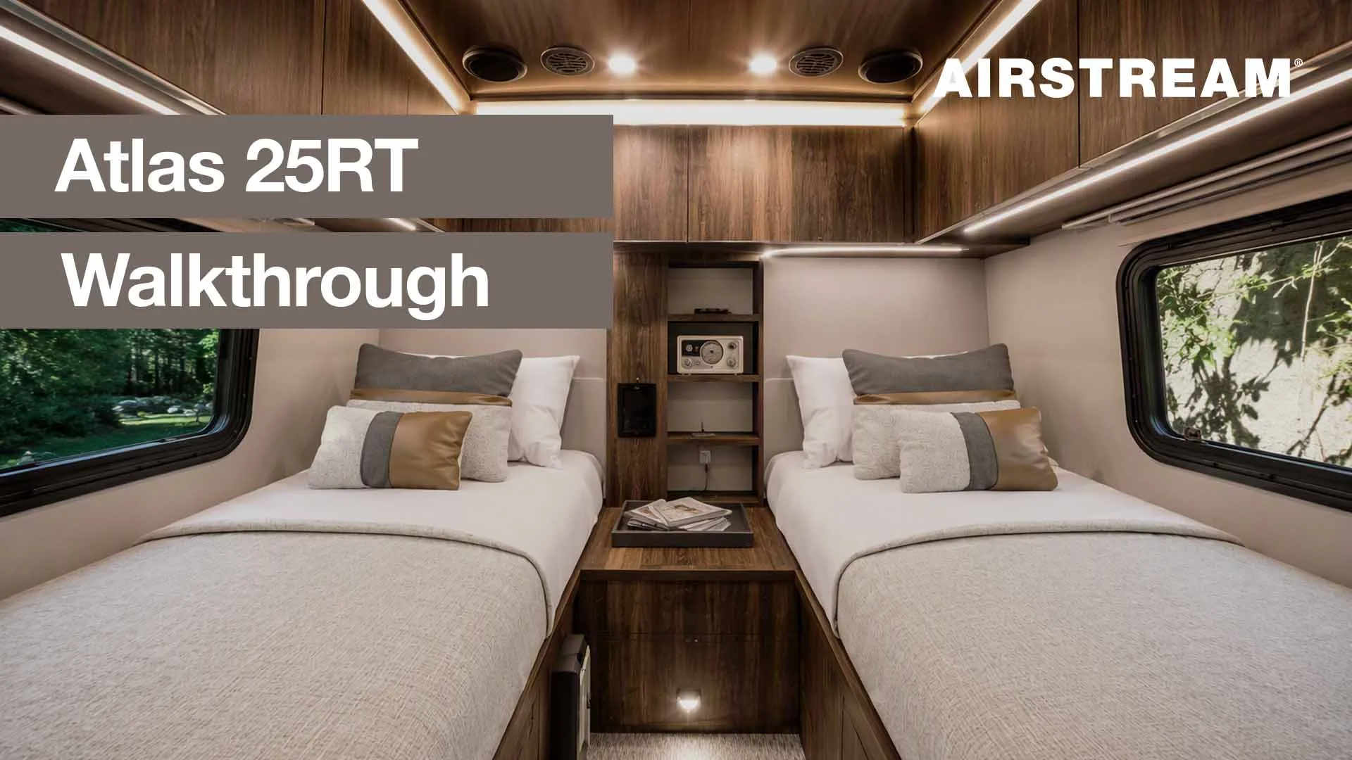Interior of a luxurious Airstream Atlas 25RT showing two twin beds with decorative pillows, wood paneling, and ambient lighting.