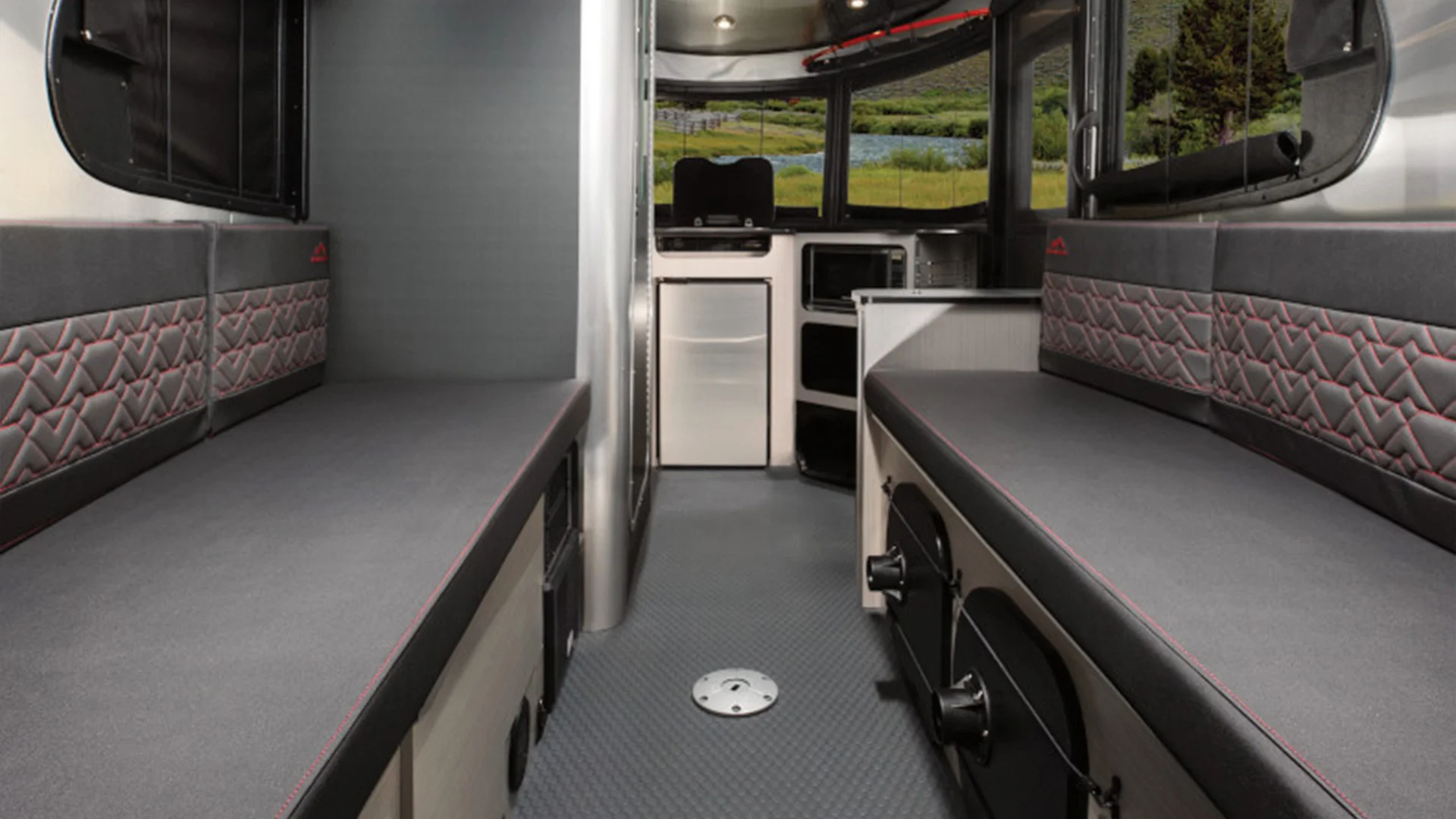 Interior of a modern camper van with two cushioned benches, storage compartments, and a kitchenette, offering a view of lush greenery outside.