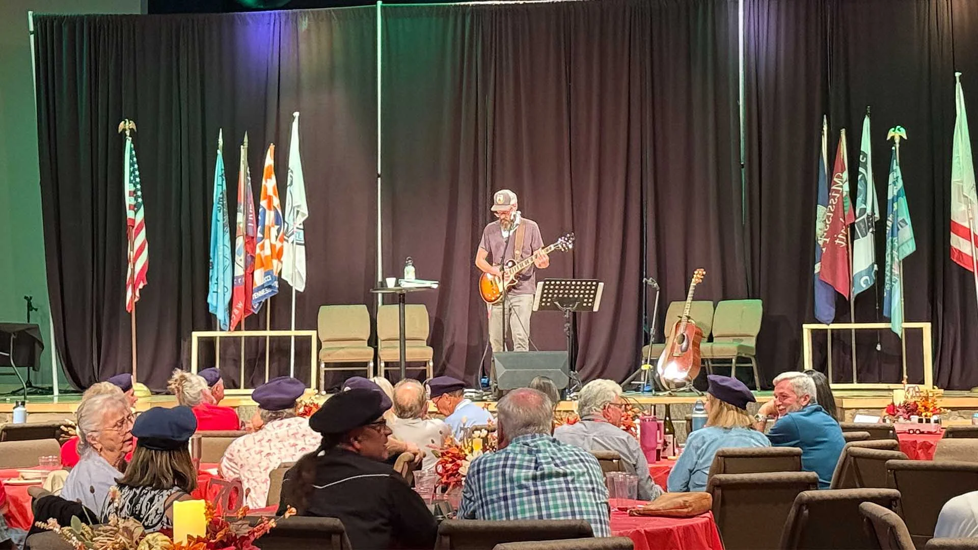 A musician performs on stage with a guitar at a Regional Airstream Club International Rally. Flags are displayed in the background, and an audience sits at tables with red tablecloths.