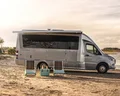 A camper van with an extended awning is parked on a beachside, with two striped chairs and a small table set up in front, under a cloudy sky.