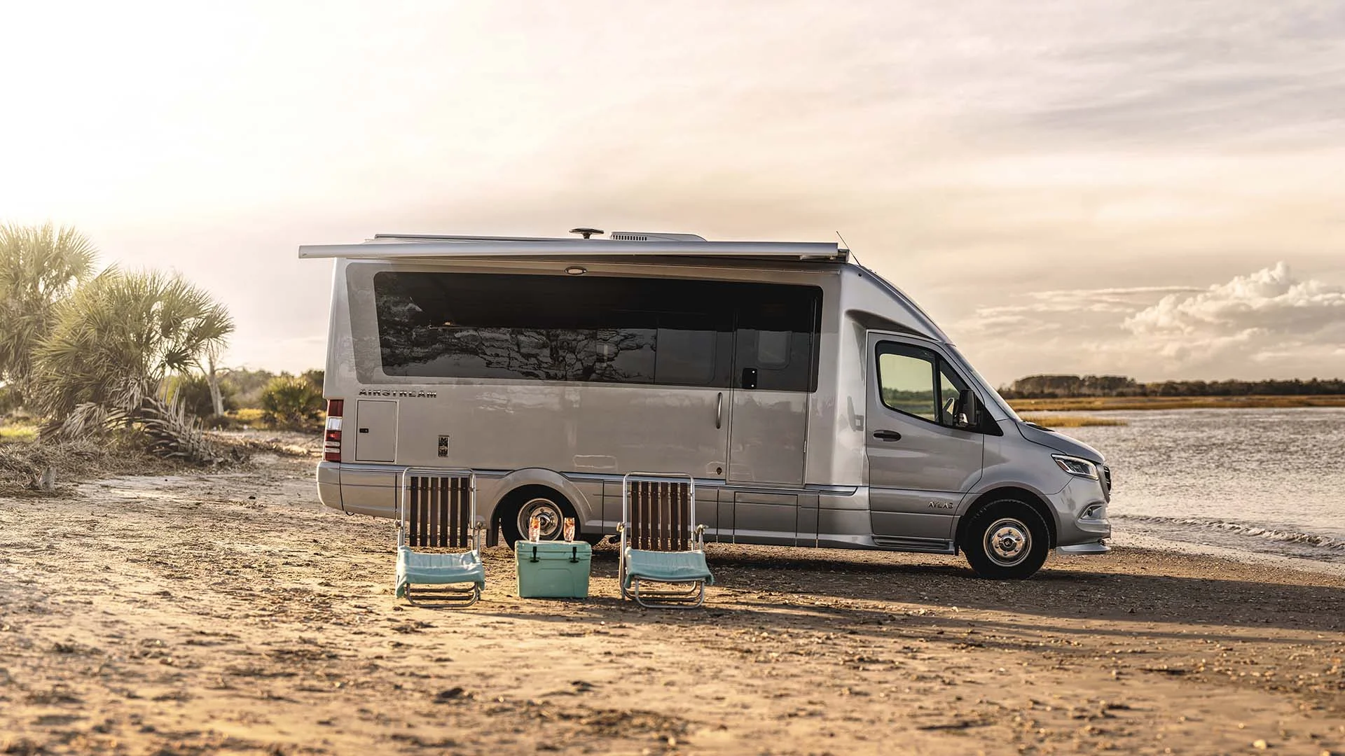 A camper van with an extended awning is parked on a beachside, with two striped chairs and a small table set up in front, under a cloudy sky.