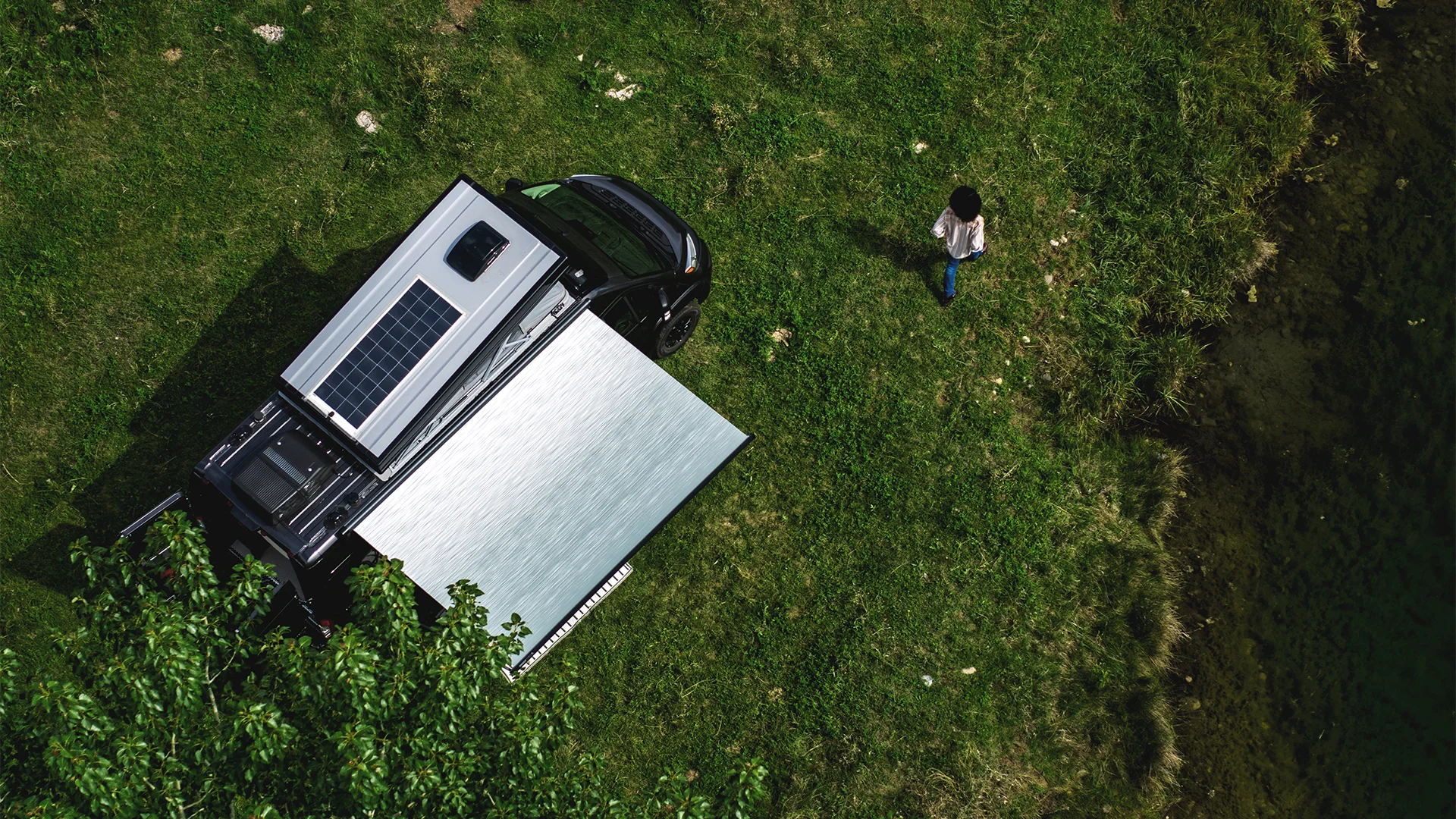 Aerial view of a black vehicle with a solar panel and awning parked on grass. A person stands nearby, with trees and a pond in the background.