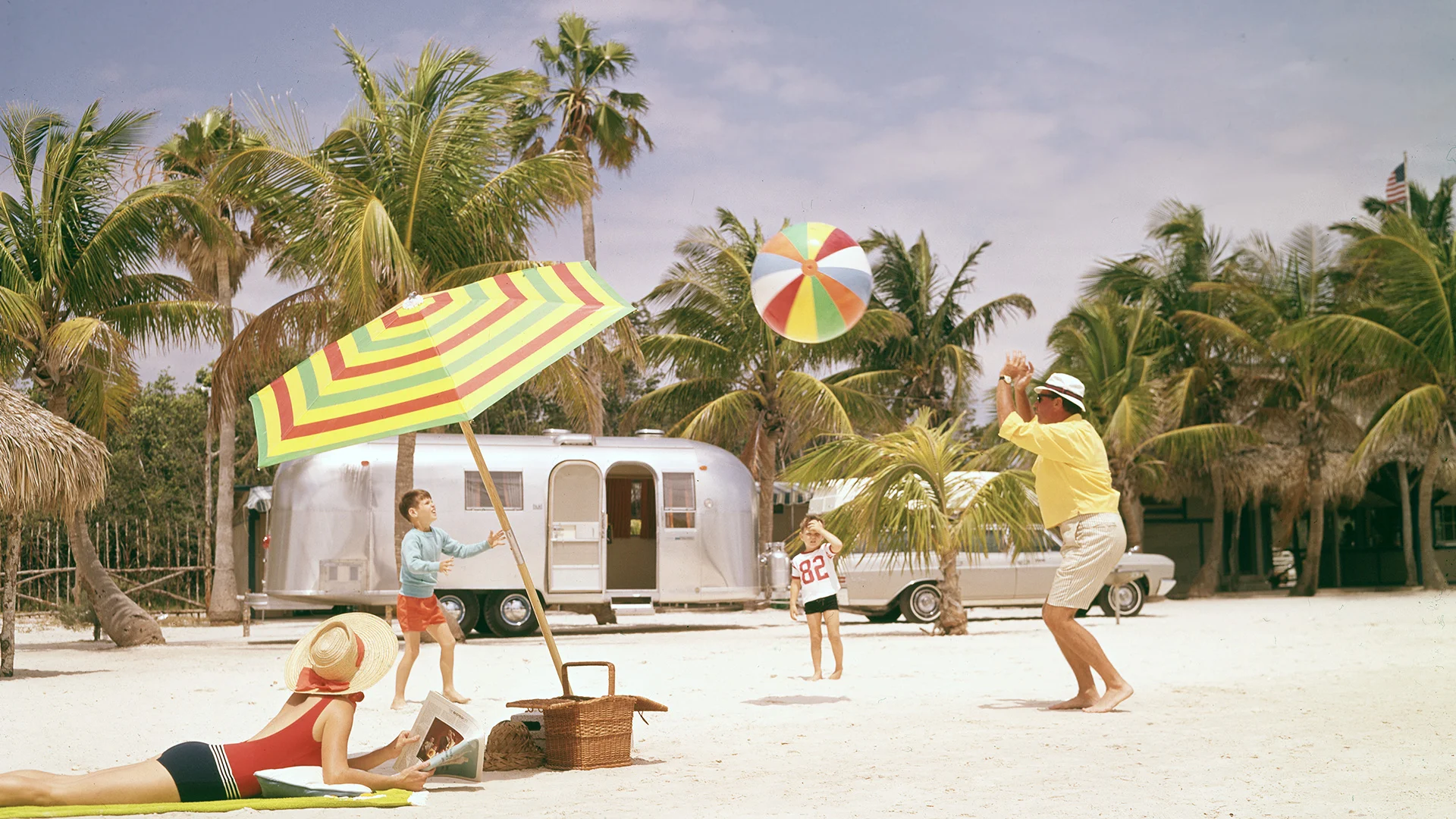 Family playing with beach ball on a sunny beach with palm trees, a striped umbrella, and a picnic basket. Silver trailer in the background.