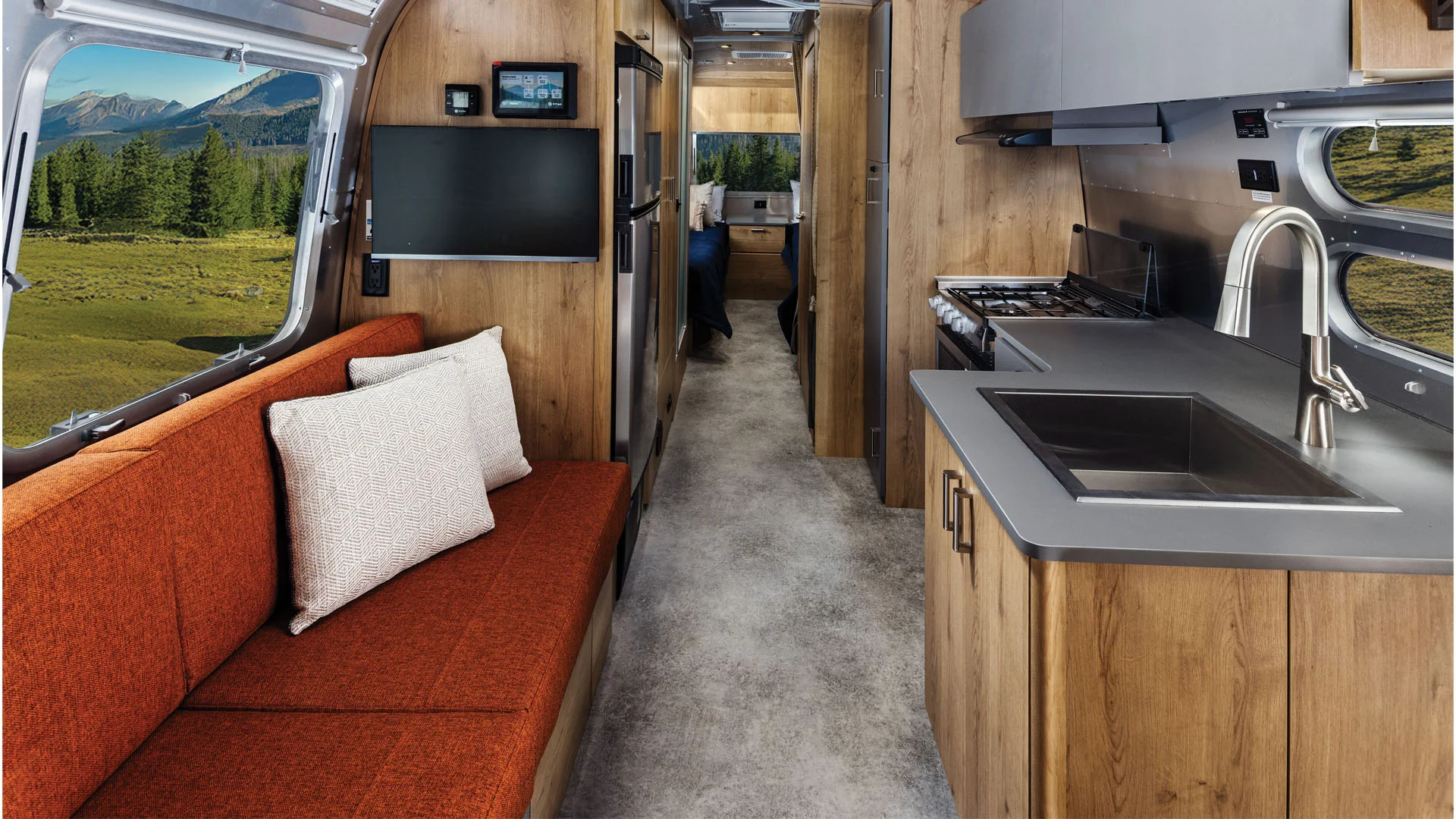 Modern RV interior with wood cabinetry, gray countertops, stainless steel sink and appliances, orange sofa with pillows, TV, and large windows showing green fields and mountains outside.