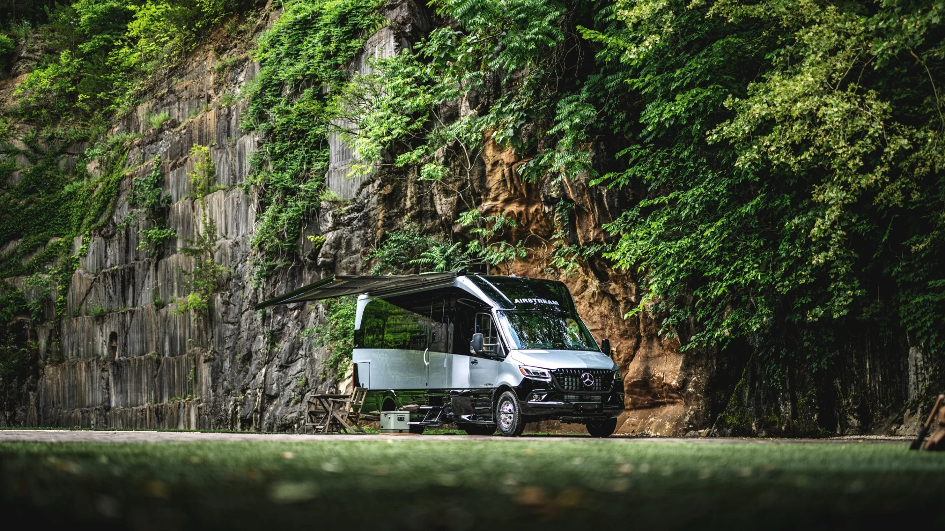 A camper van with an awning extended, parked near a rocky cliffside covered in lush greenery.