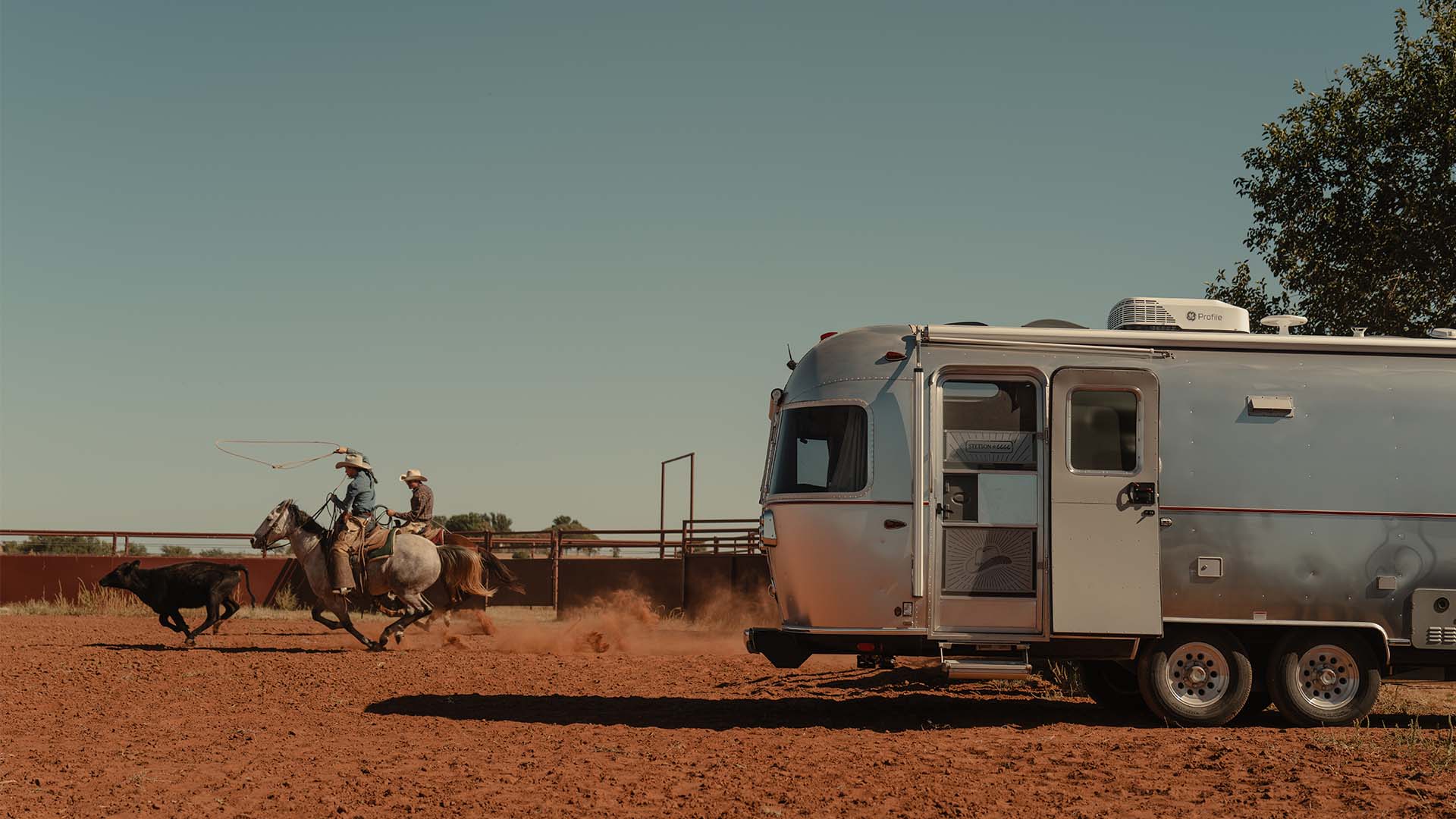 Two cowboys on horses lasso a calf in a dusty arena beside a shiny silver trailer under a clear sky.