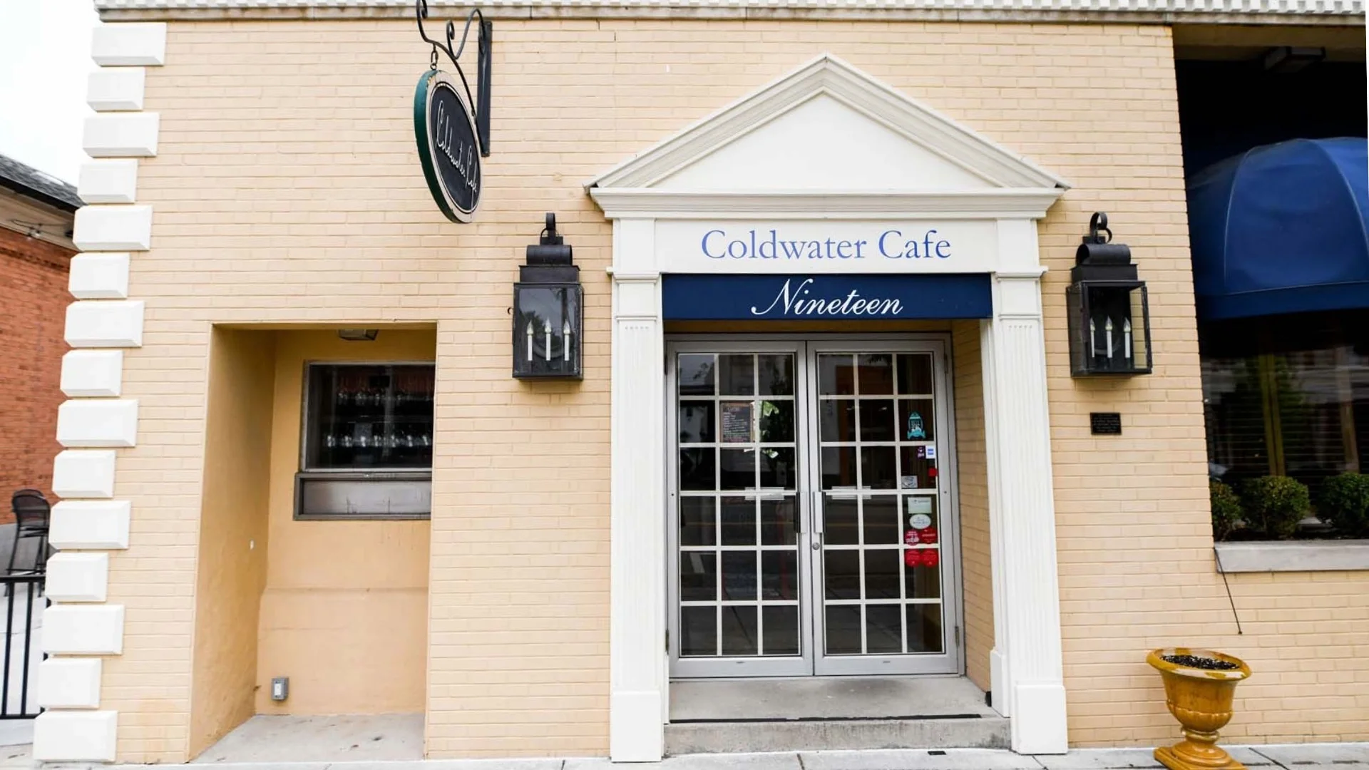Entrance of Coldwater Cafe Nineteen, featuring a light brick facade, two lanterns, and a blue awning with white text.