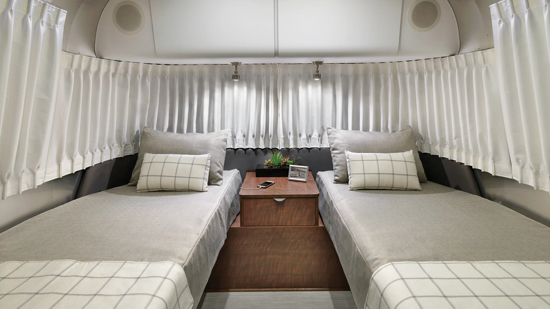 Interior of a cozy RV bedroom with two twin beds, gray bedding, checkered cushions, wooden nightstand with phone and plant, and white curtains.
