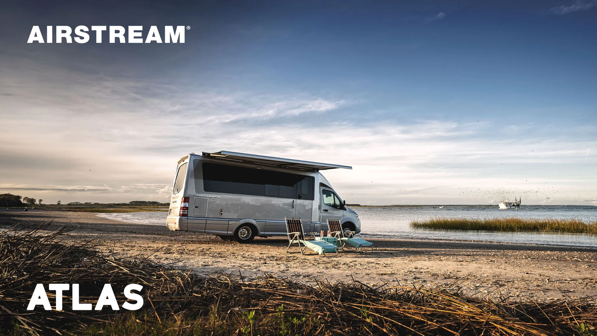 A silver Airstream Atlas RV parked on a beach with two lounge chairs facing the water under a partly cloudy sky.