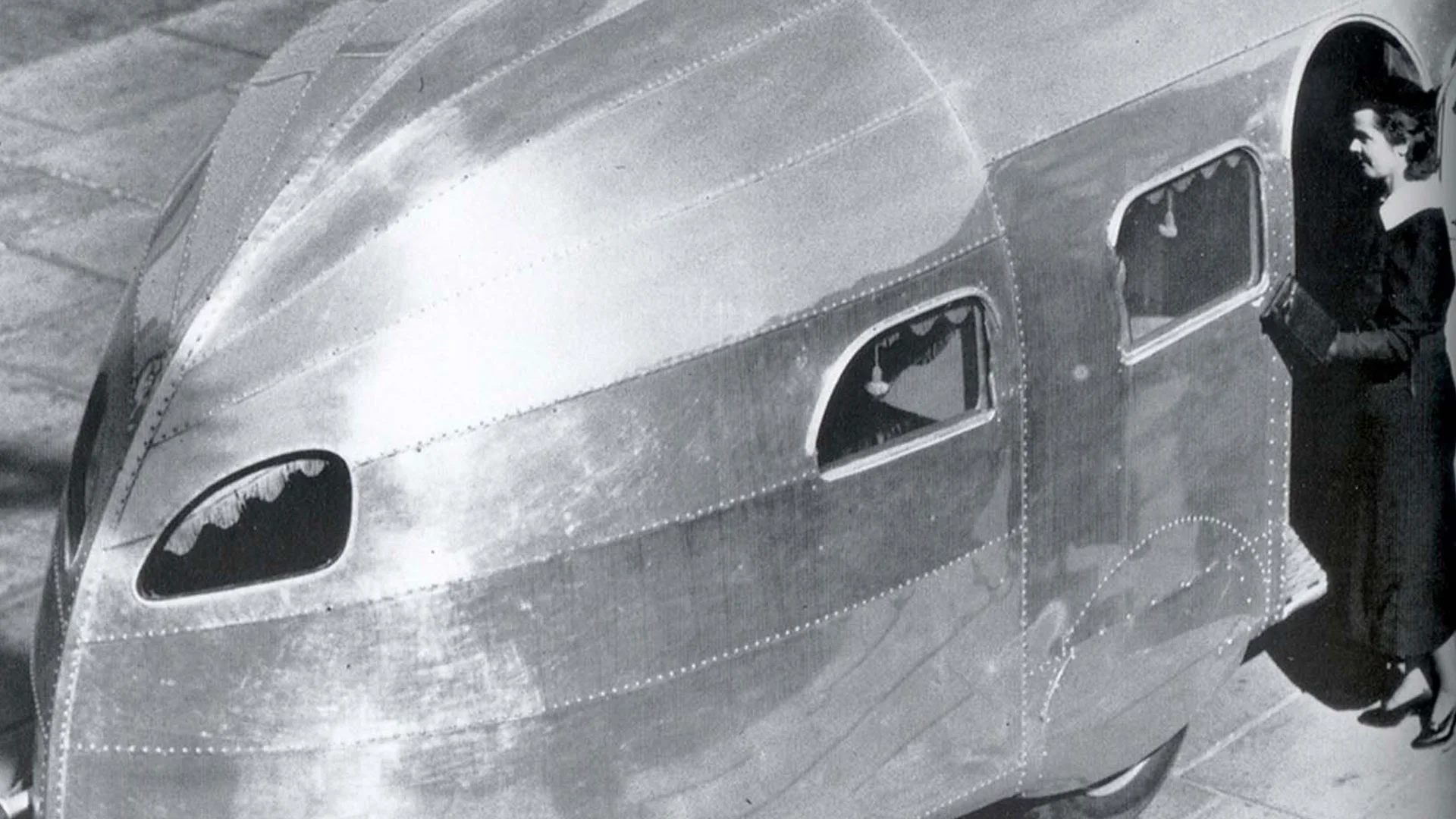 A woman standing beside a sleek, vintage, metallic trailer with rounded windows and a futuristic design.