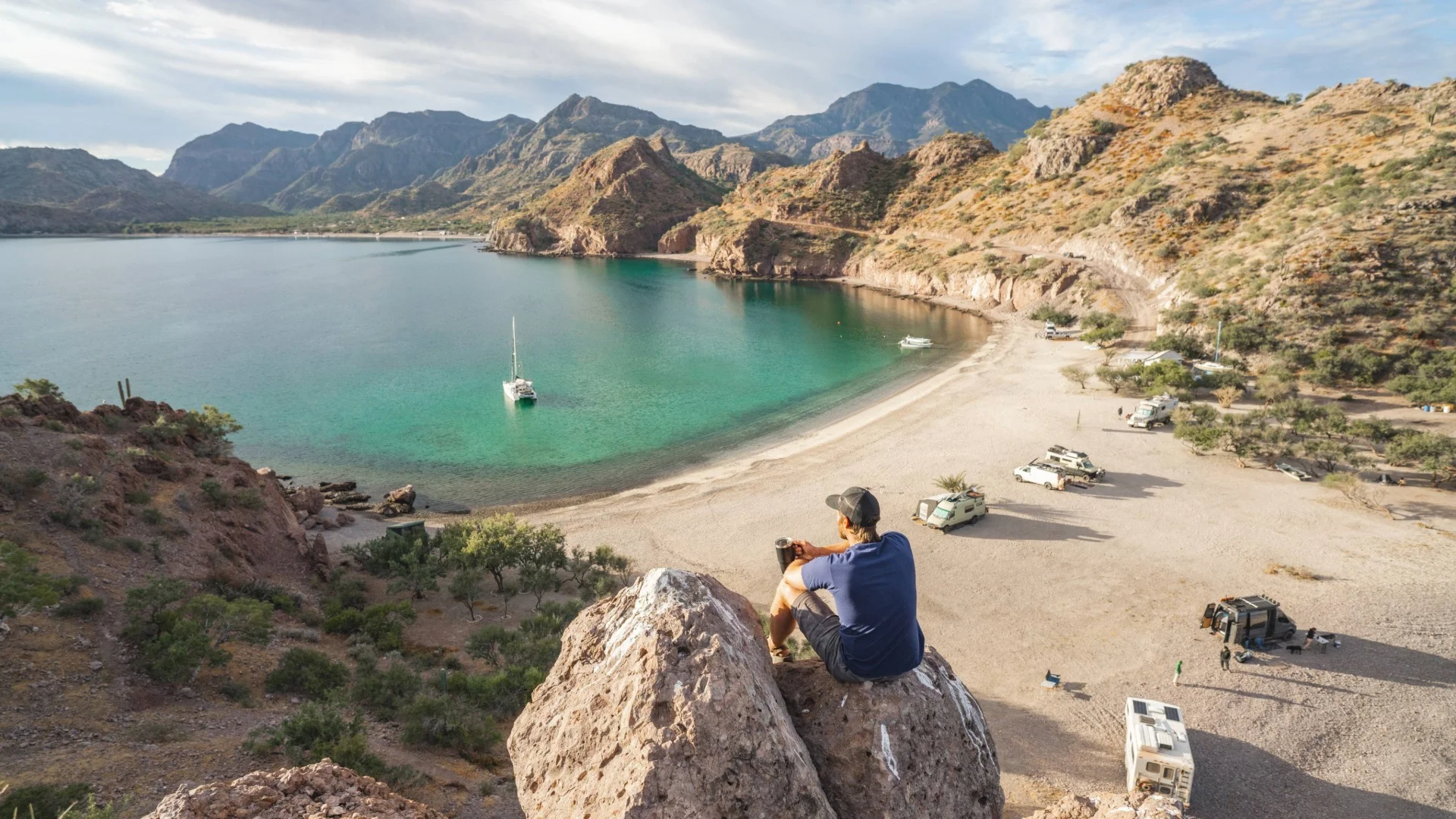 Person sits on rock overlooking a beach with parked vehicles, a sailboat on turquoise water, and rugged mountains in the background.