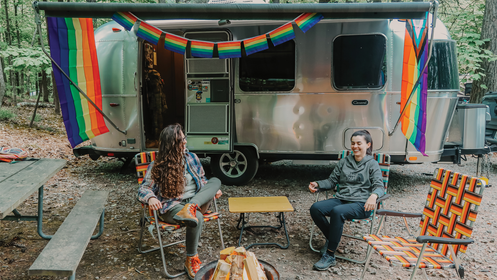 Two people relax in foldable chairs outside a camper van with rainbow decorations in a forest campsite.