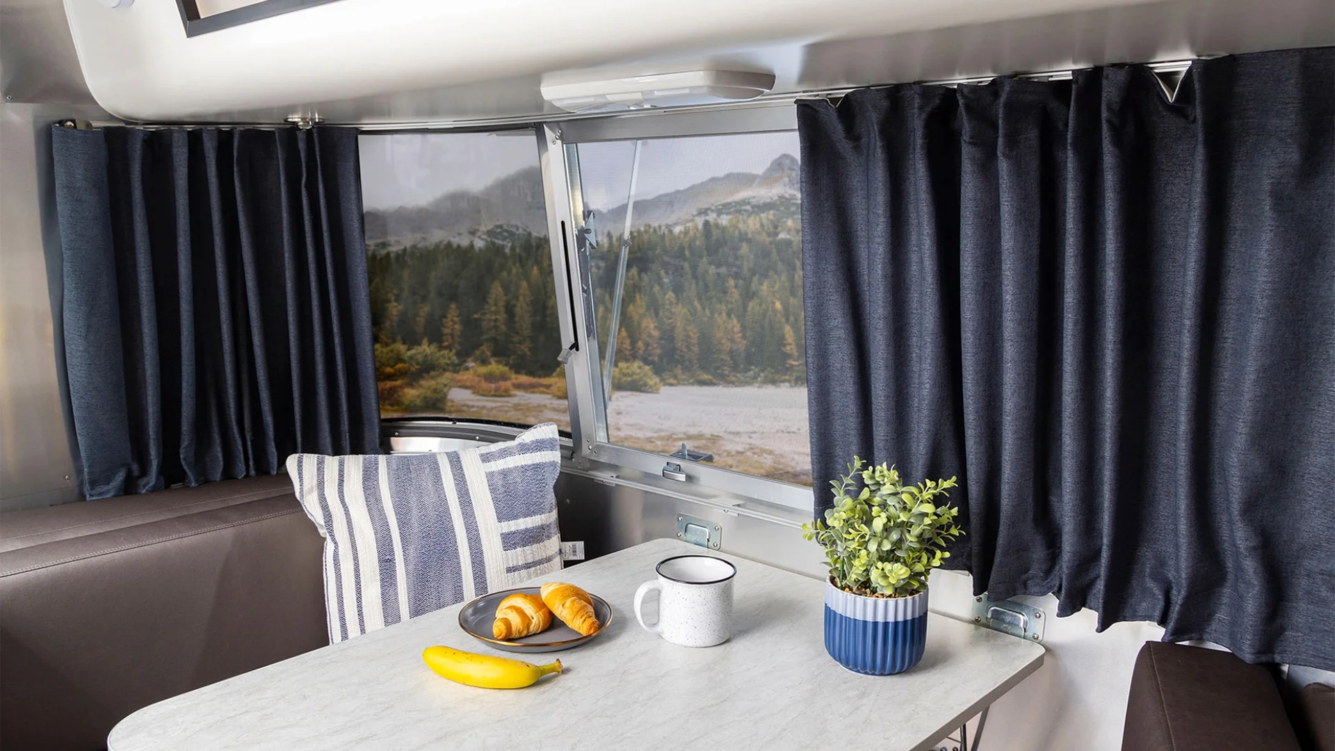RV interior with dark curtains, a table set with a banana, croissants, a mug, a cushion, and a potted plant by a window showing a forest view.