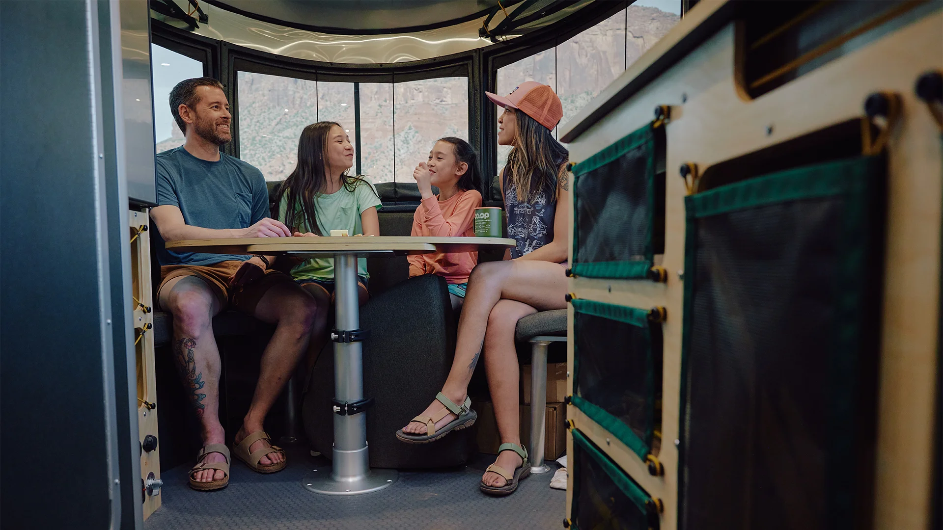 A family of four sits around a table, smiling inside a cozy camper with large windows.