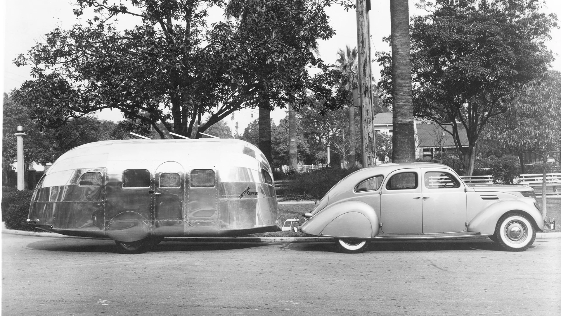 A vintage car tows a shiny, metallic camper trailer on a street lined with trees. The car has a streamlined design typical of the mid-20th century.