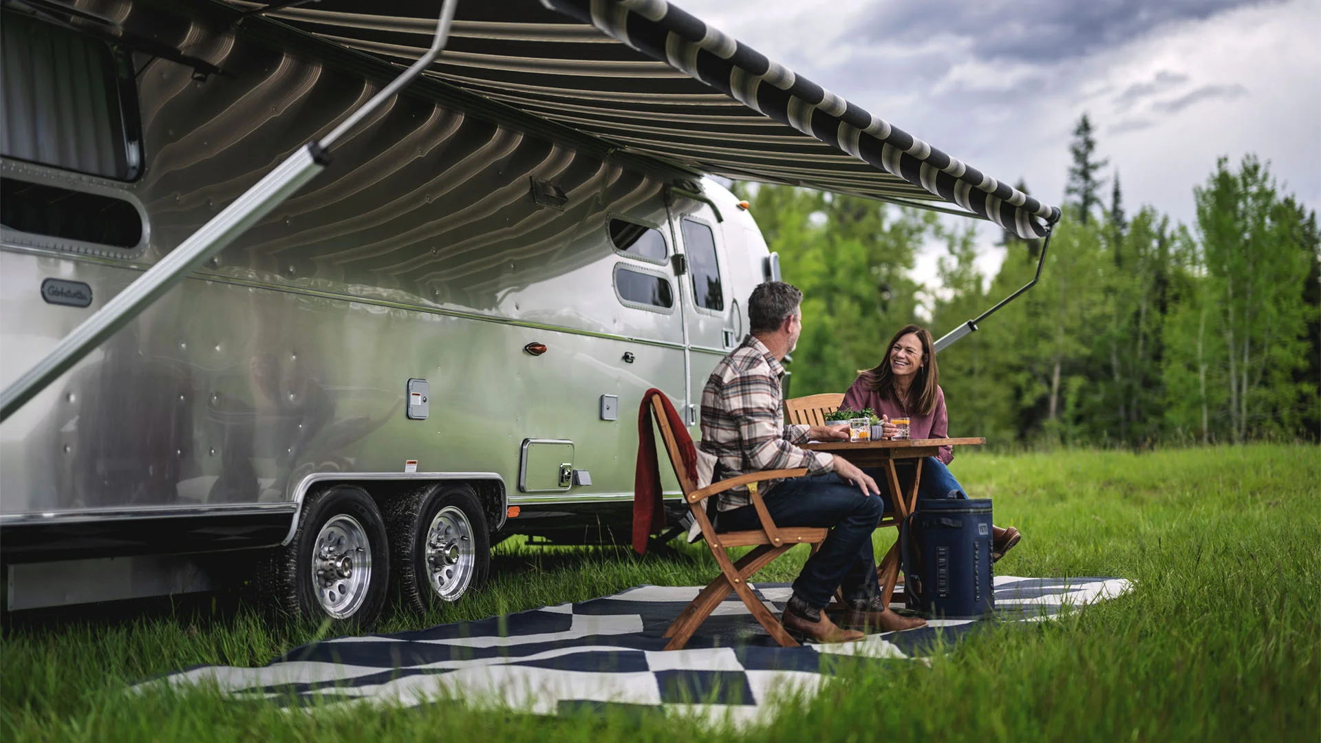 A man and woman sit at a table under the awning of a silver camper trailer, enjoying food and drinks on a picnic mat in a grassy field with trees in the background.