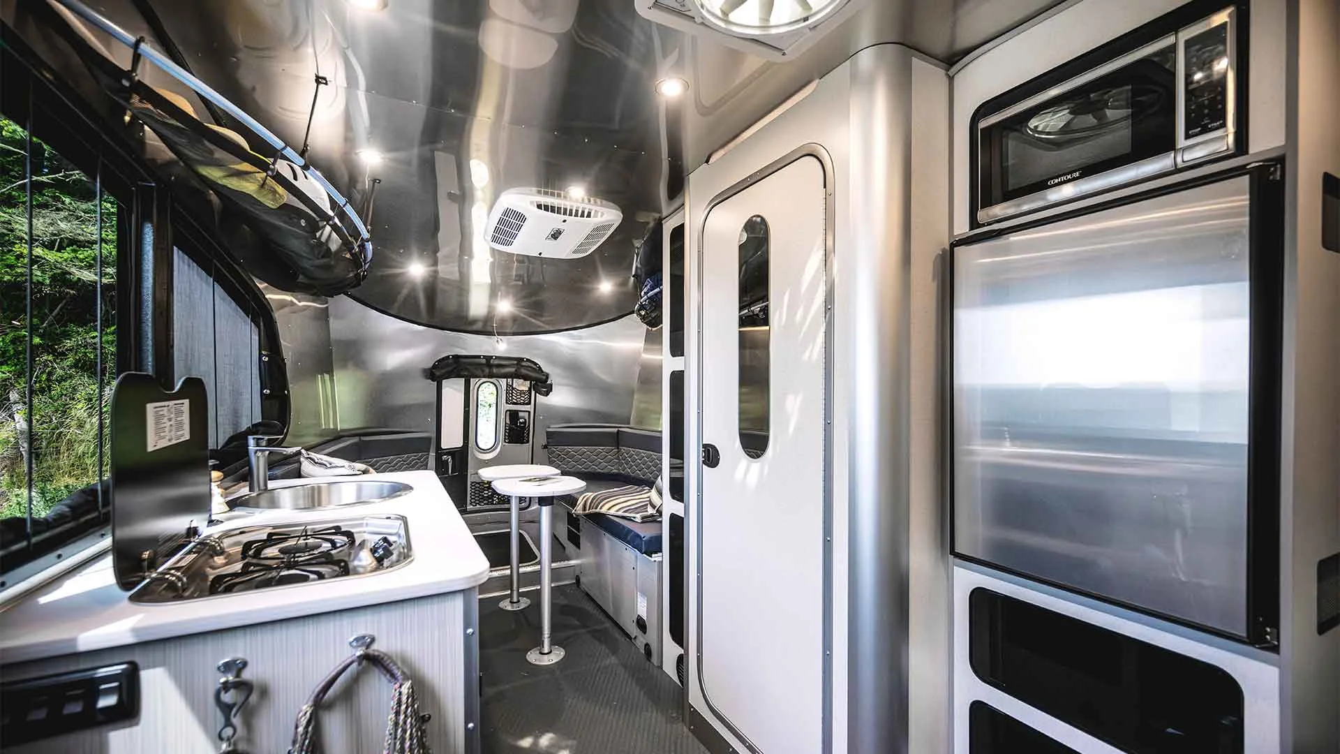 Modern trailer interior with a compact kitchen, stove, and fridge on the right. Small seating area and table towards the back. Sleek, metallic design.