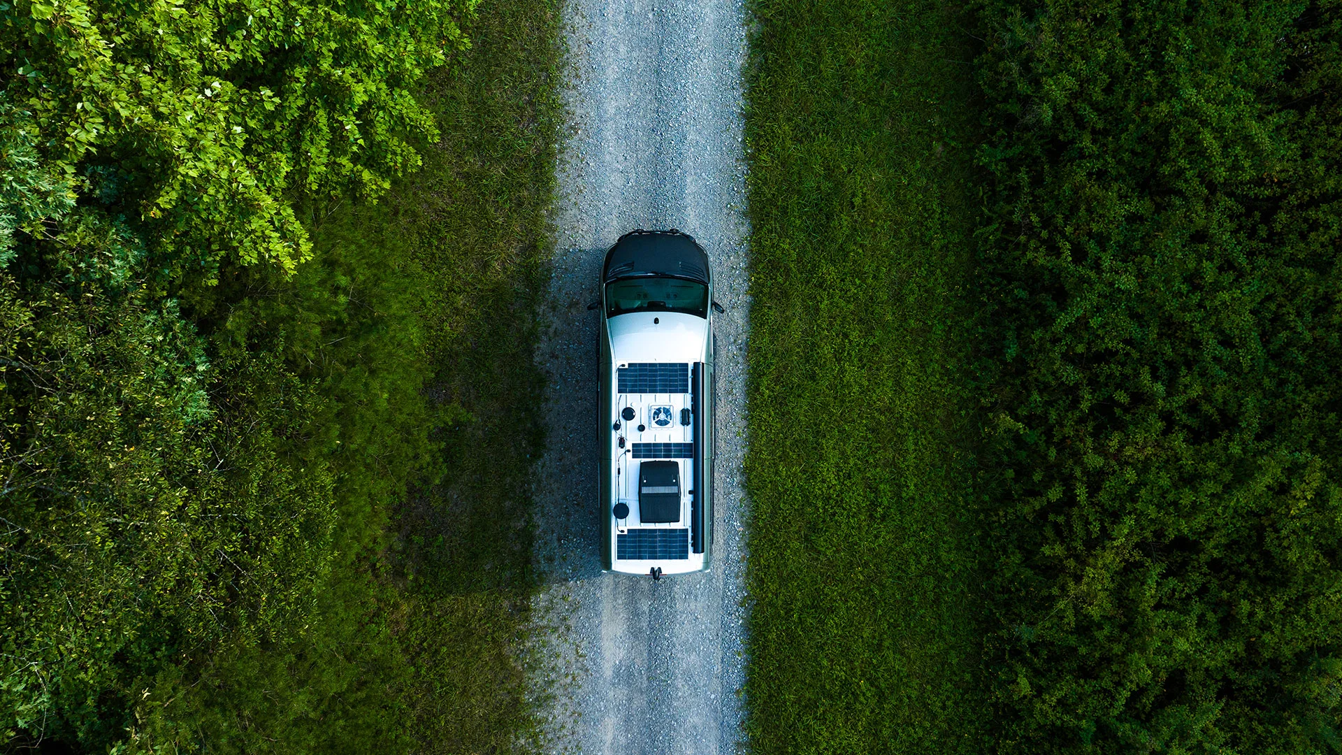 Aerial view of a camper van with solar panels on a gravel road, surrounded by dense green vegetation.
