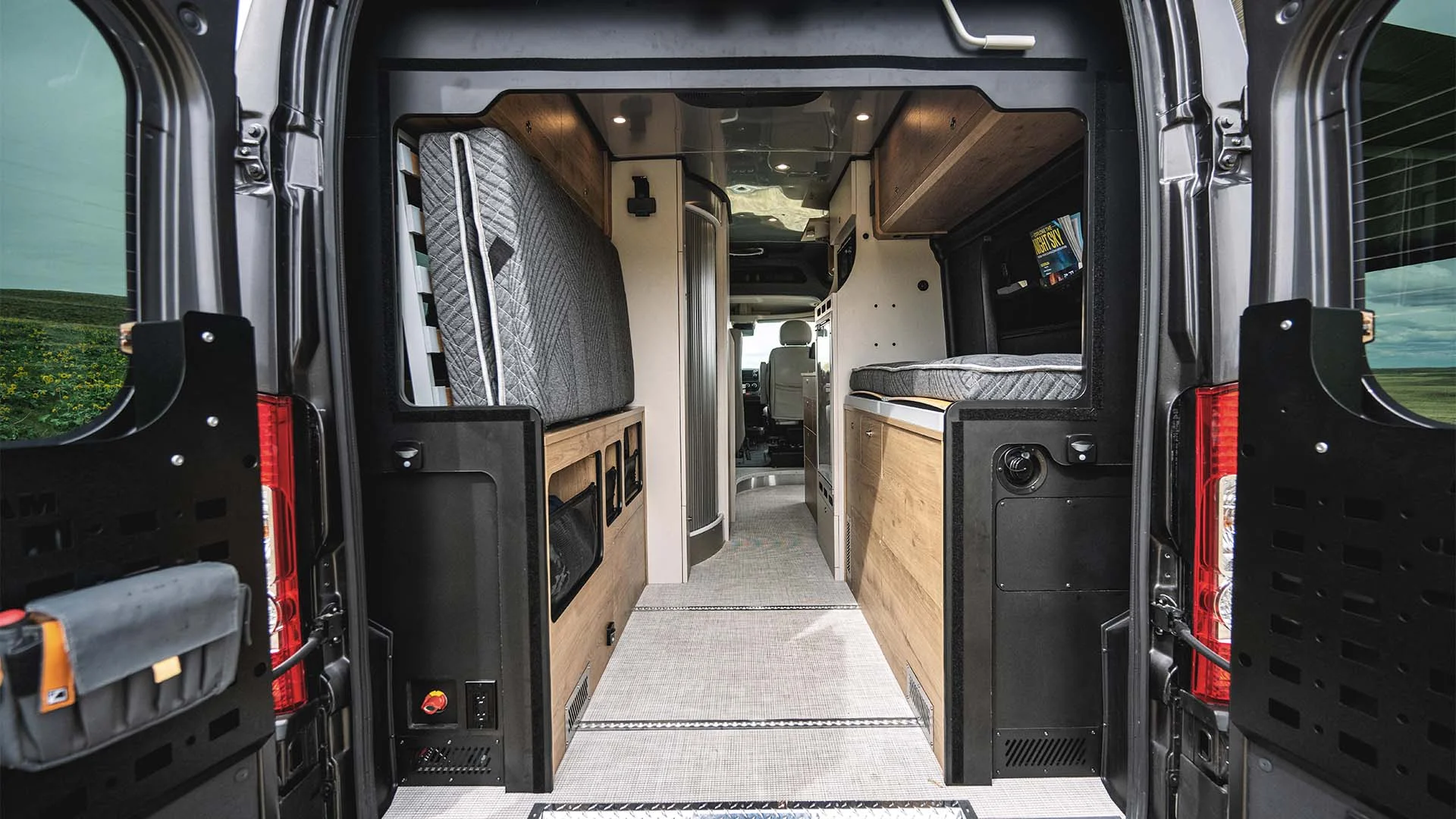 Interior view of a camper van with a bed, cabinets, and a compact aisle leading to the driver's area, seen from the rear doors.