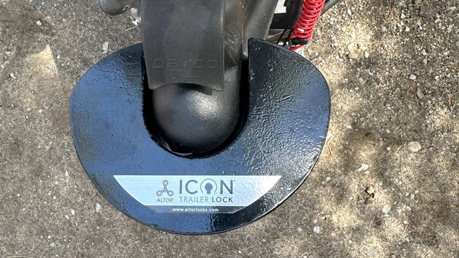 Close-up of an Altor ICON trailer lock securing a trailer hitch, with a textured black surface and branding on a metal plate.