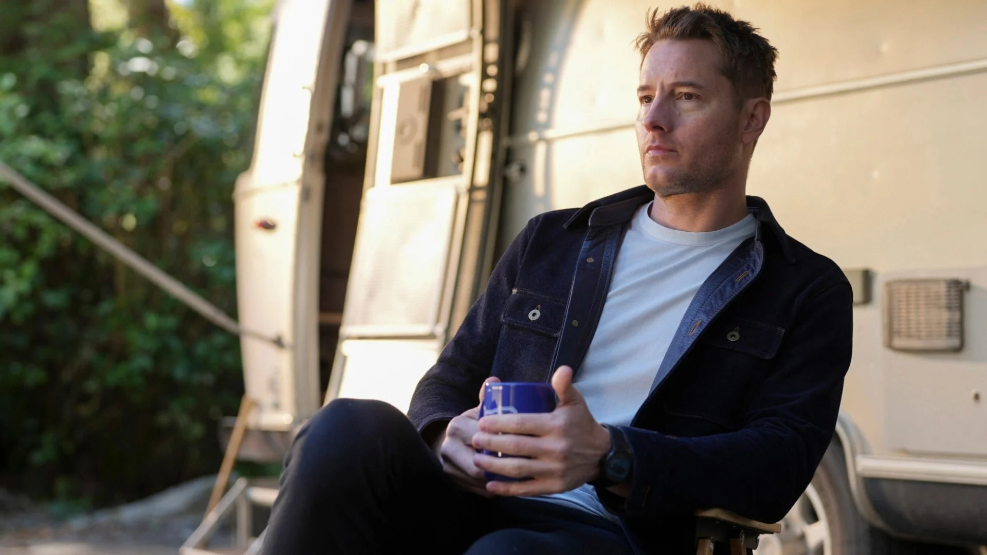 Man sitting outside a trailer, holding a blue mug, wearing a dark jacket and white shirt, with greenery in the background.