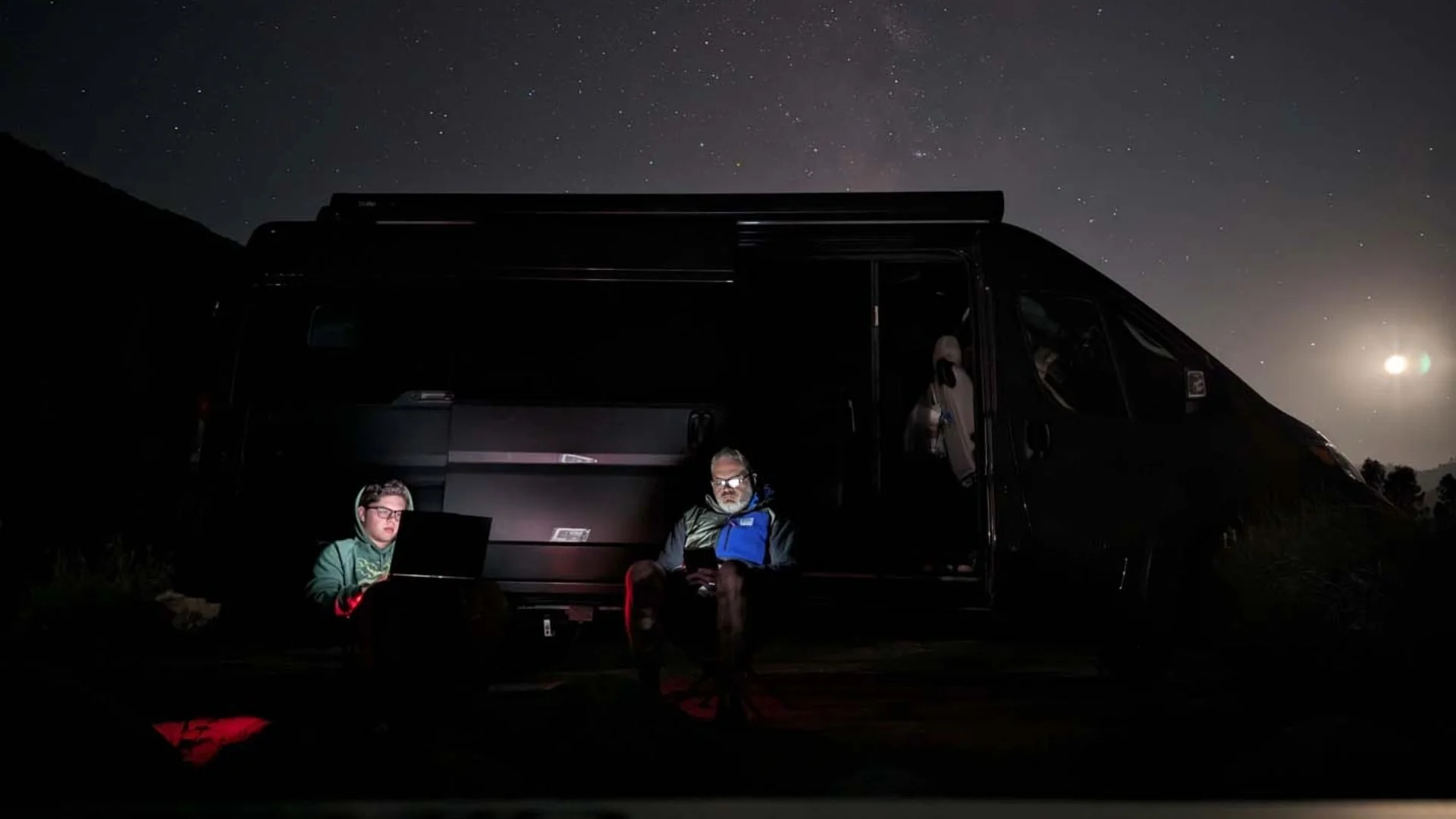 Two people sitting outside a van at night, lit by laptop screens, with a starry sky and moon in the background.