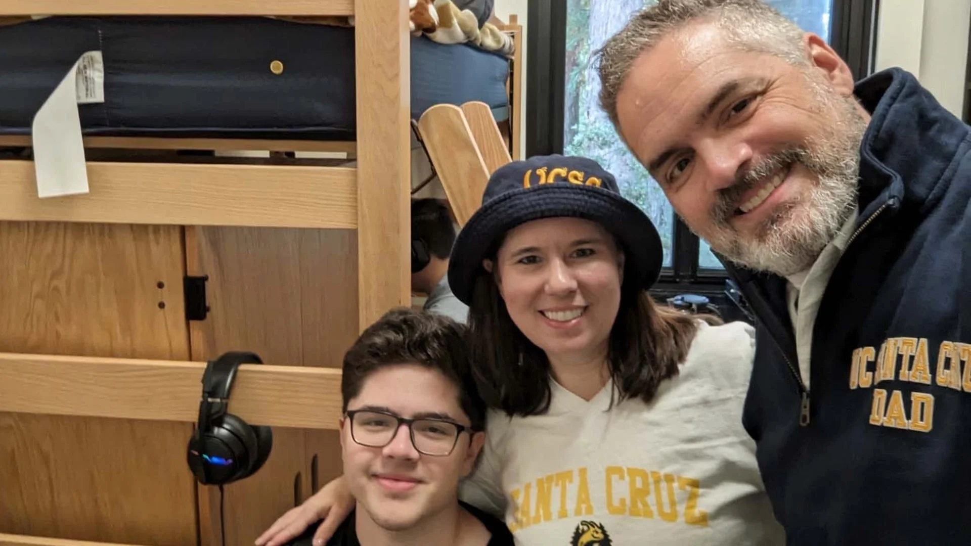 Three people smiling in a dorm room. A young man sits with two adults standing behind him, all wearing UCSC attire. A bunk bed is visible.