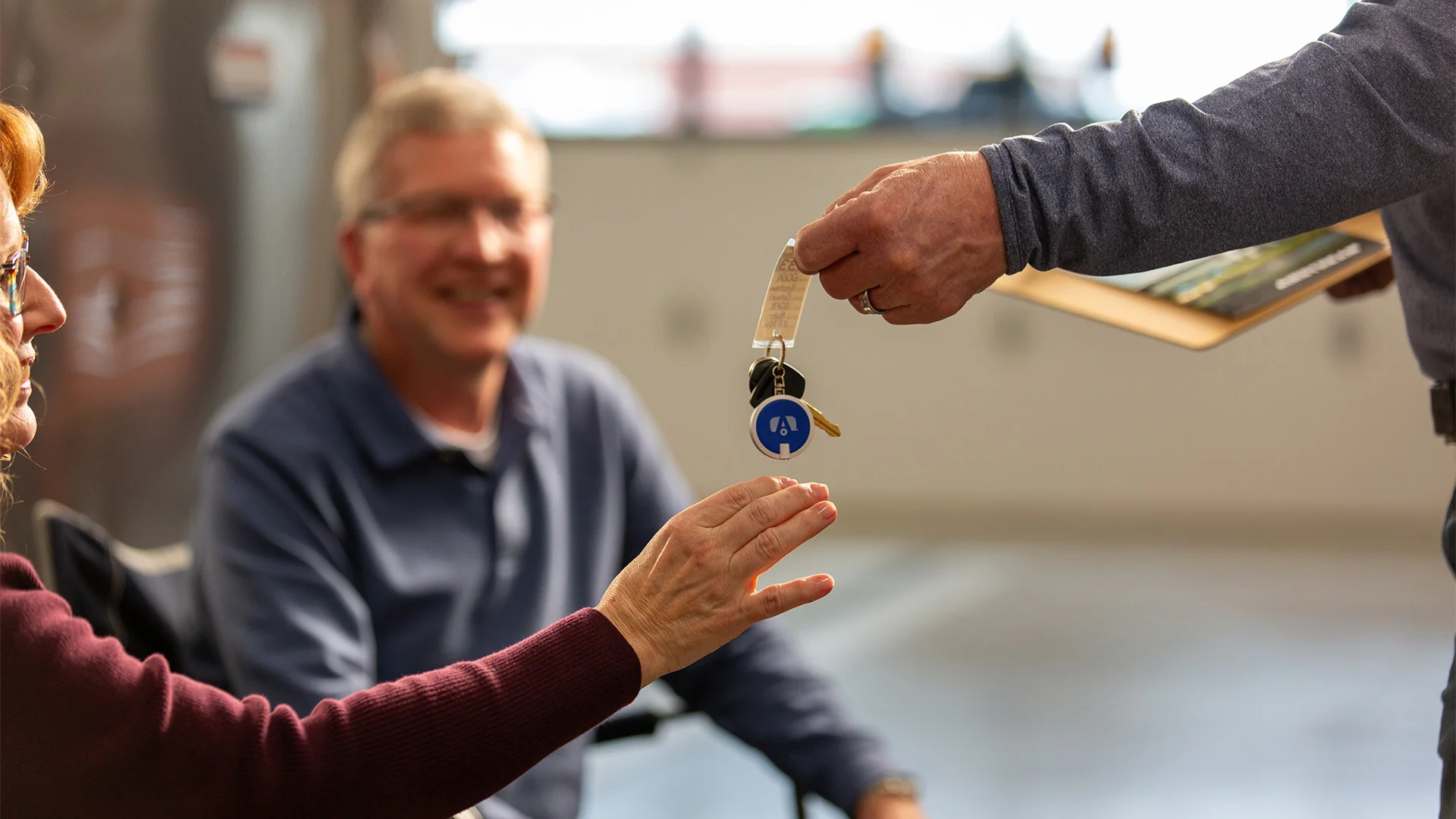 A person hands over keys to another person who is seated, while another seated person looks on with a smile.