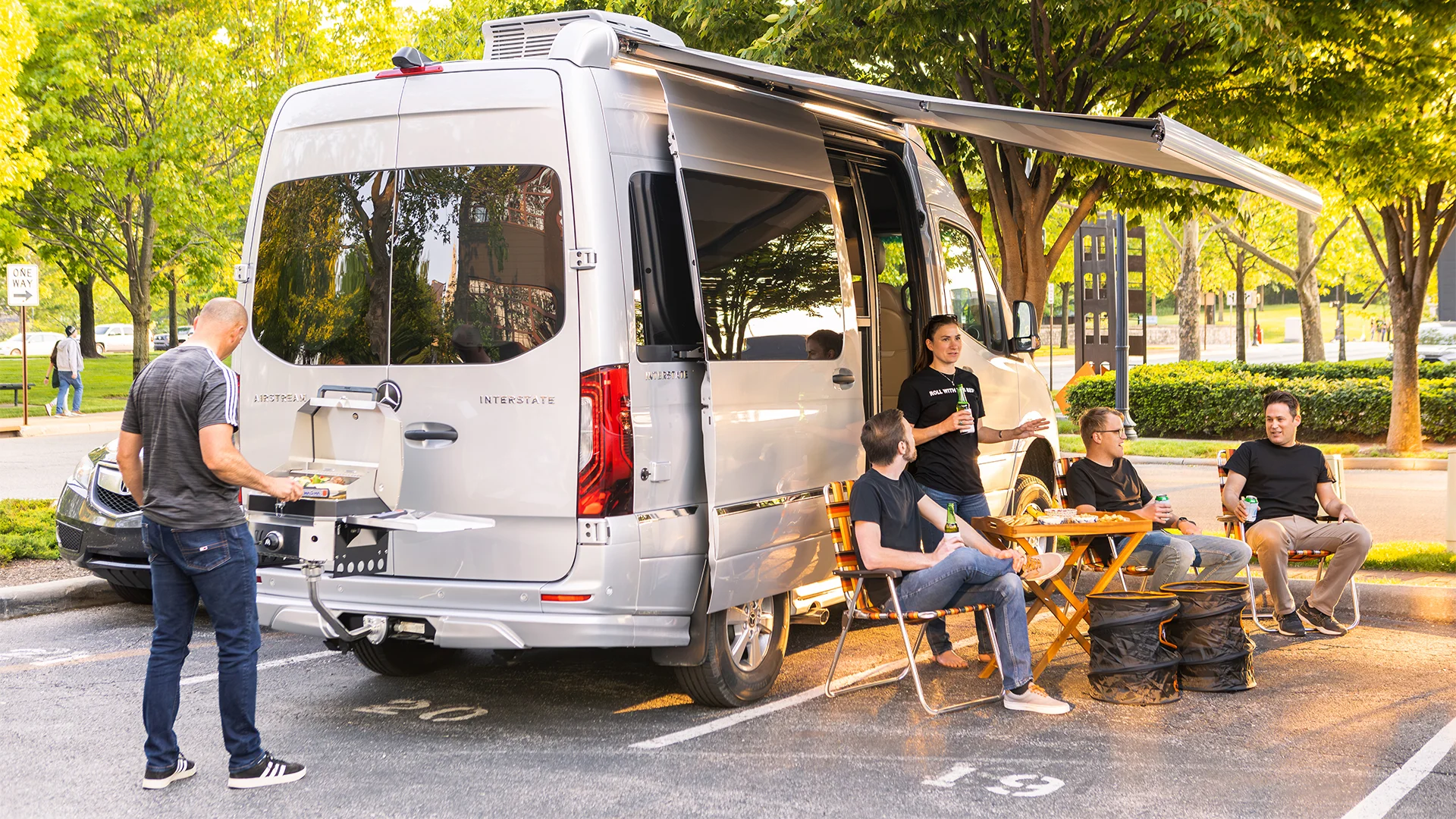 A group of people gathered around a camper van with an open awning, sitting and chatting. One person grills food on a built-in outdoor cooker.