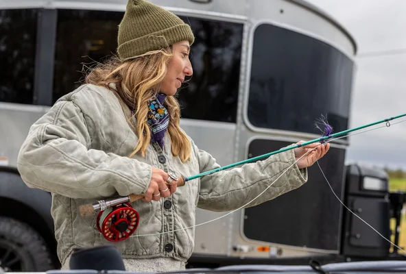 A woman in a green beanie and gray jacket prepares a fishing rod outdoors, standing beside a silver trailer with a black tire visible in the background.