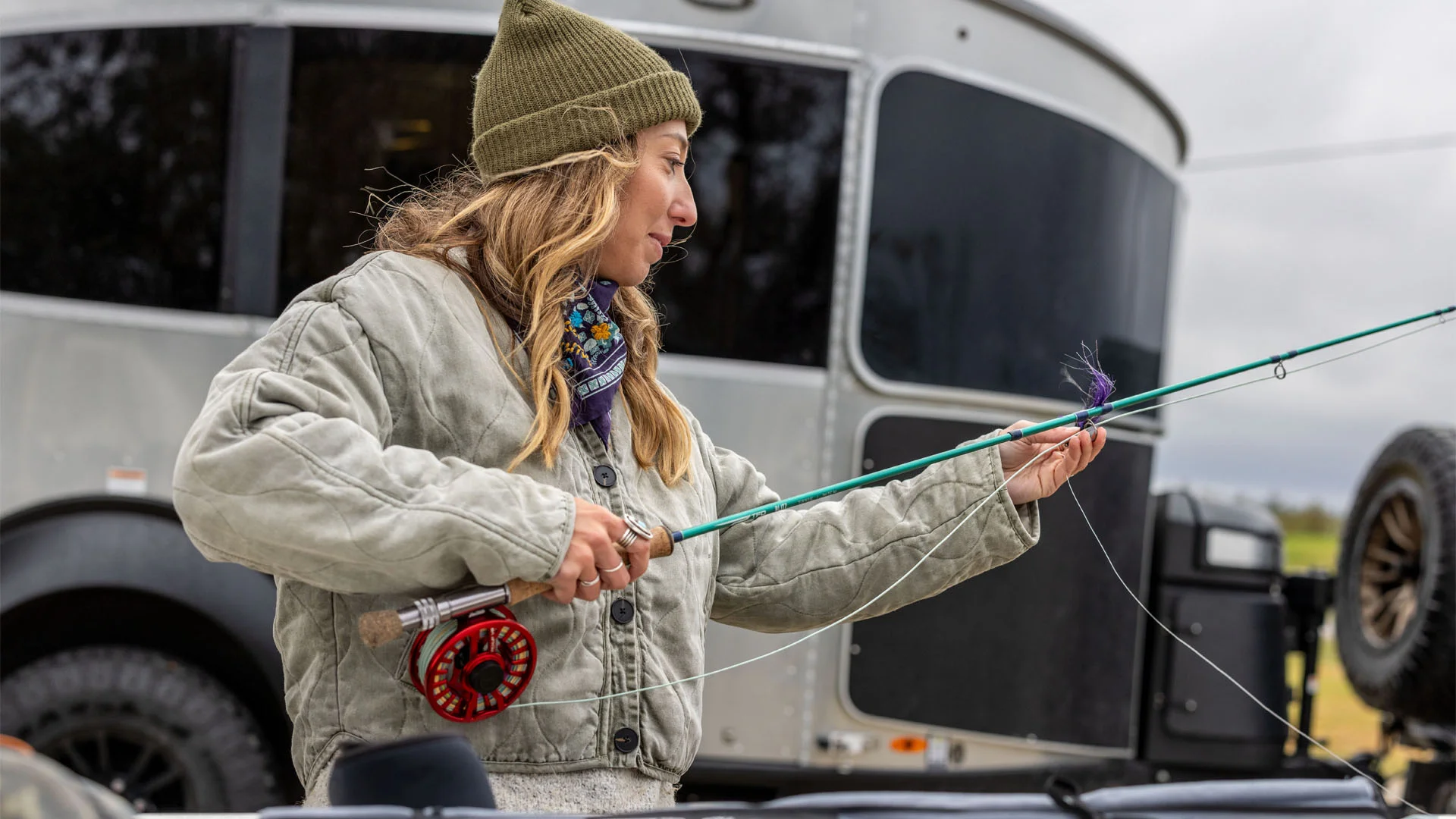 A woman in a green beanie and gray jacket prepares a fishing rod outdoors, standing beside a silver trailer with a black tire visible in the background.