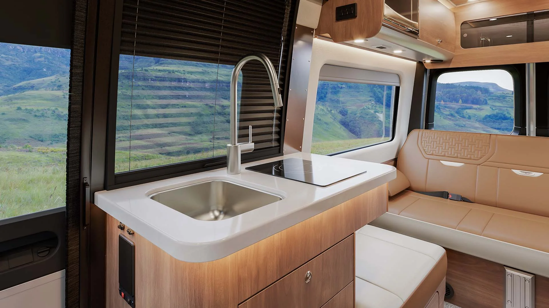 Modern camper van interior with sink, stovetop, and seating, featuring large windows offering a scenic view of rolling green hills.