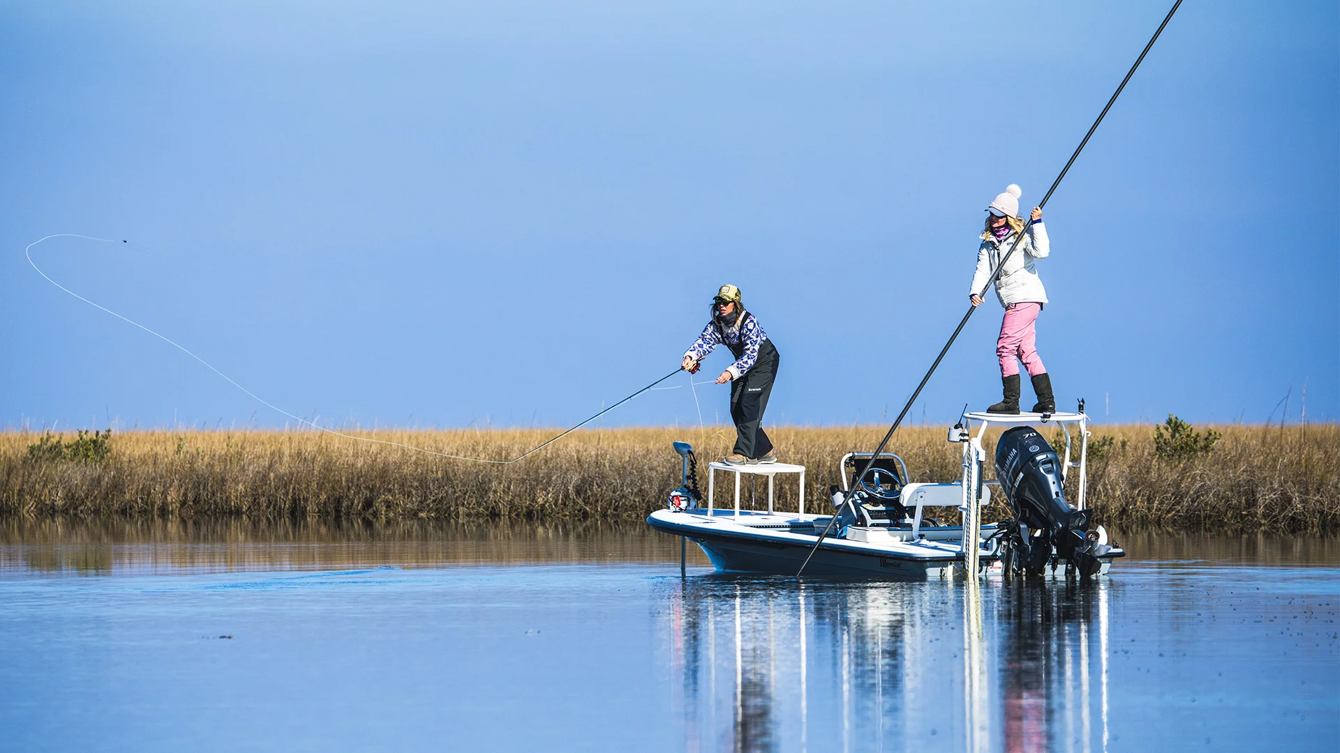 Two people fishing from a boat in a calm marsh, with one casting a line and the other standing on a raised platform steering with a pole.
