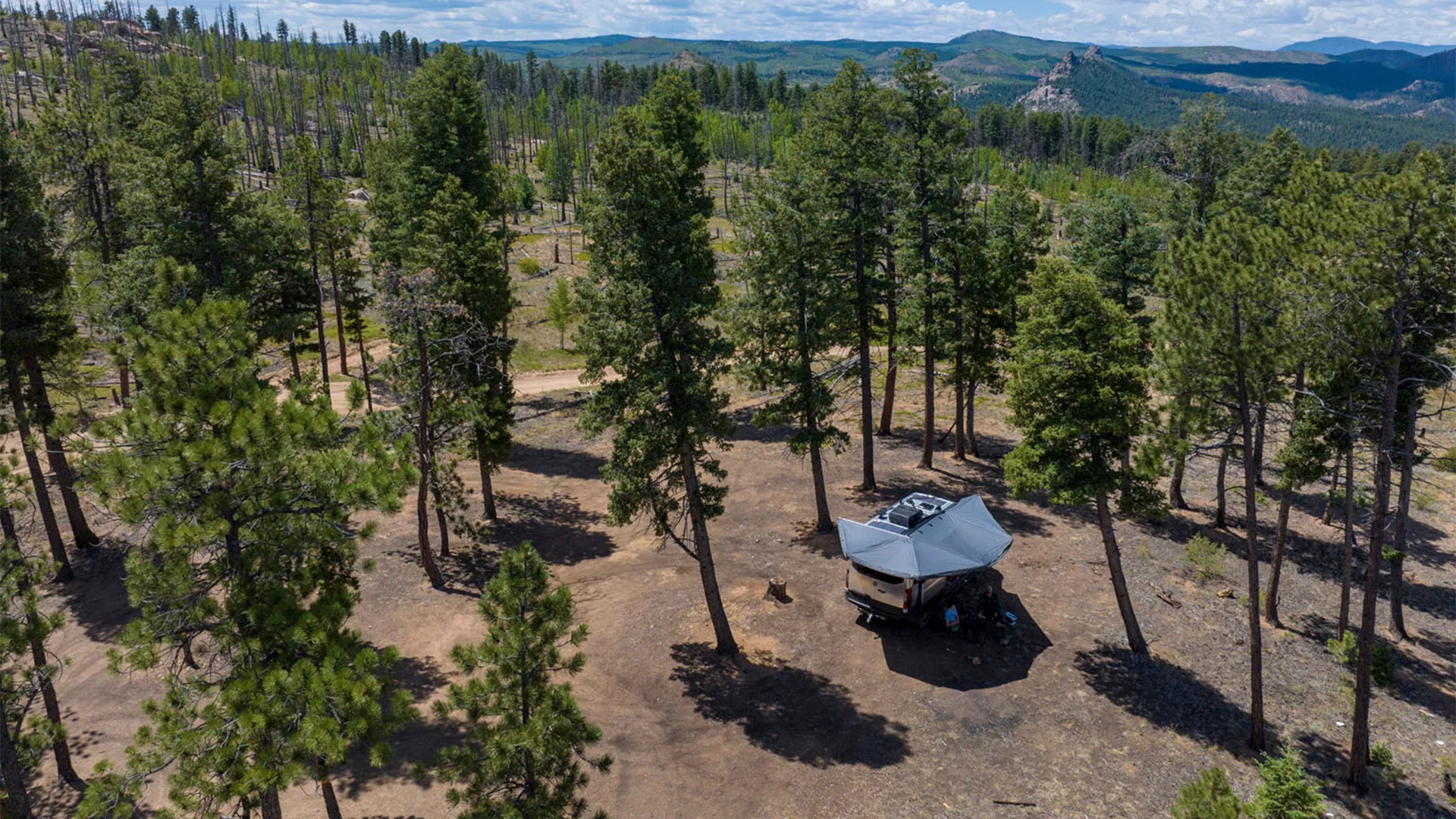 A camper with an awning is set up in a sparse, wooded area surrounded by tall trees, with mountainous terrain in the distance under a blue sky.