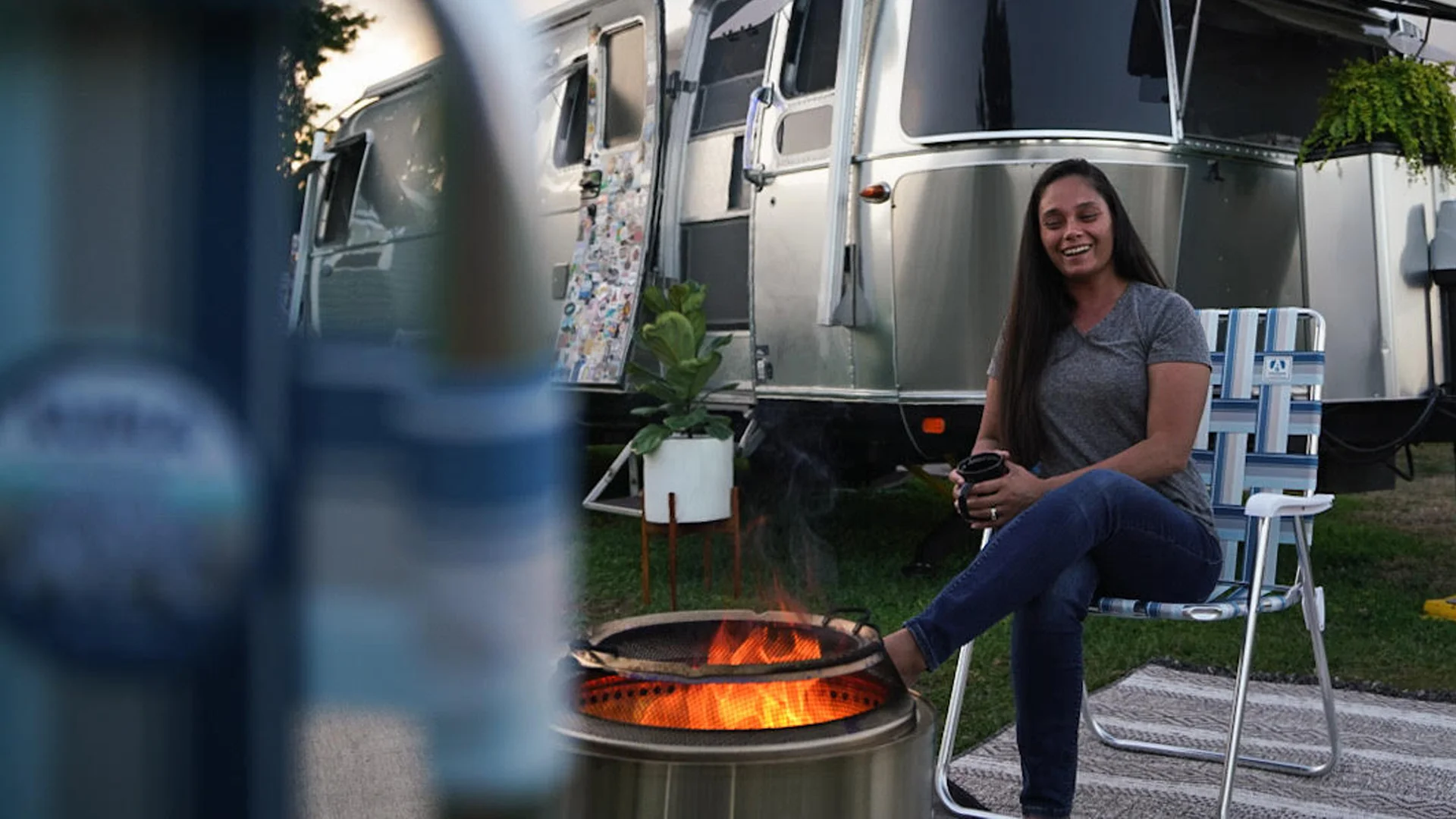 A woman sits on a lawn chair by a fire pit, smiling and holding a mug, with a silver Airstream trailer and plants in the background at a campsite.