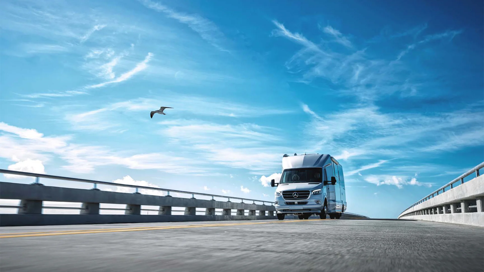 A silver van drives on a bridge under a clear blue sky with wispy clouds; a bird flies overhead.