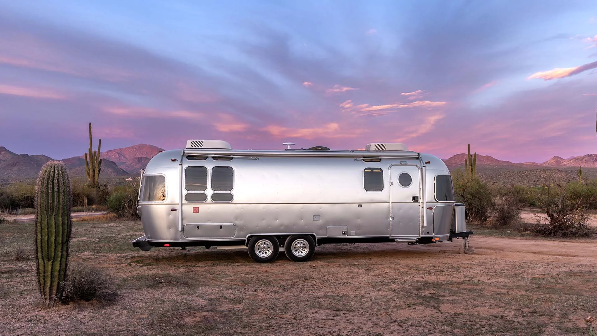 Silver Airstream trailer parked in a desert landscape at sunset, surrounded by cacti and mountains under a colorful sky.