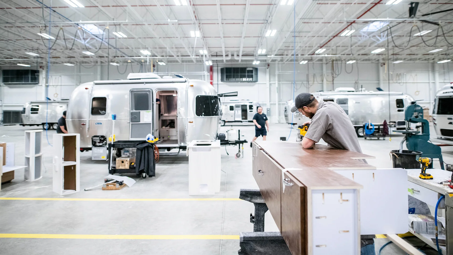 Worker assembling furniture in a large factory with several shiny Airstream trailers in the background.