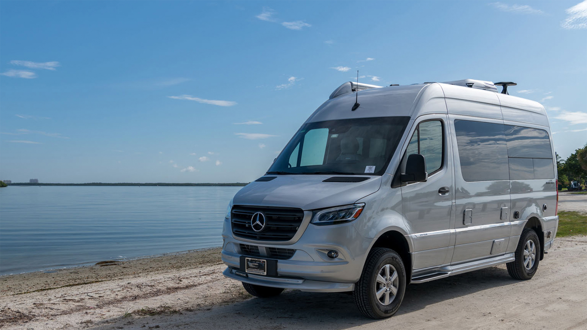 Silver Mercedes-Benz camper van parked near a calm beach with a clear blue sky in the background.