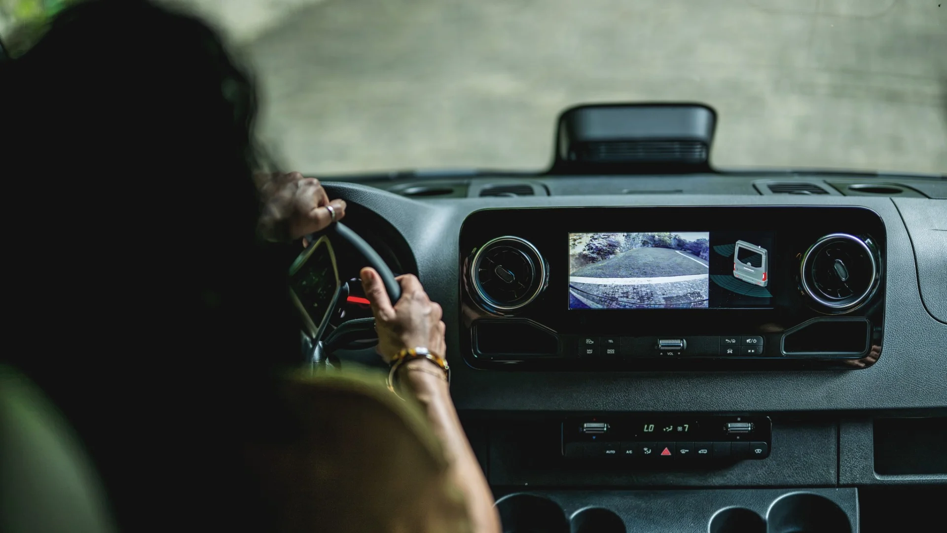 Person driving a vehicle, holding the steering wheel with both hands. The dashboard screen displays a backup camera view.