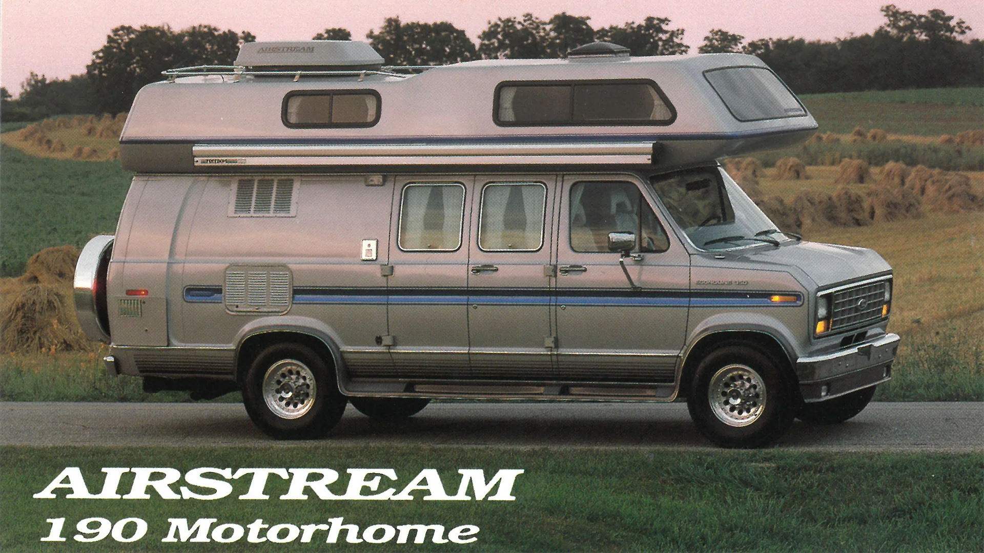 Silver Airstream 190 motorhome parked on a rural road, featuring a high roof and vintage design elements, with hay bales in the background.