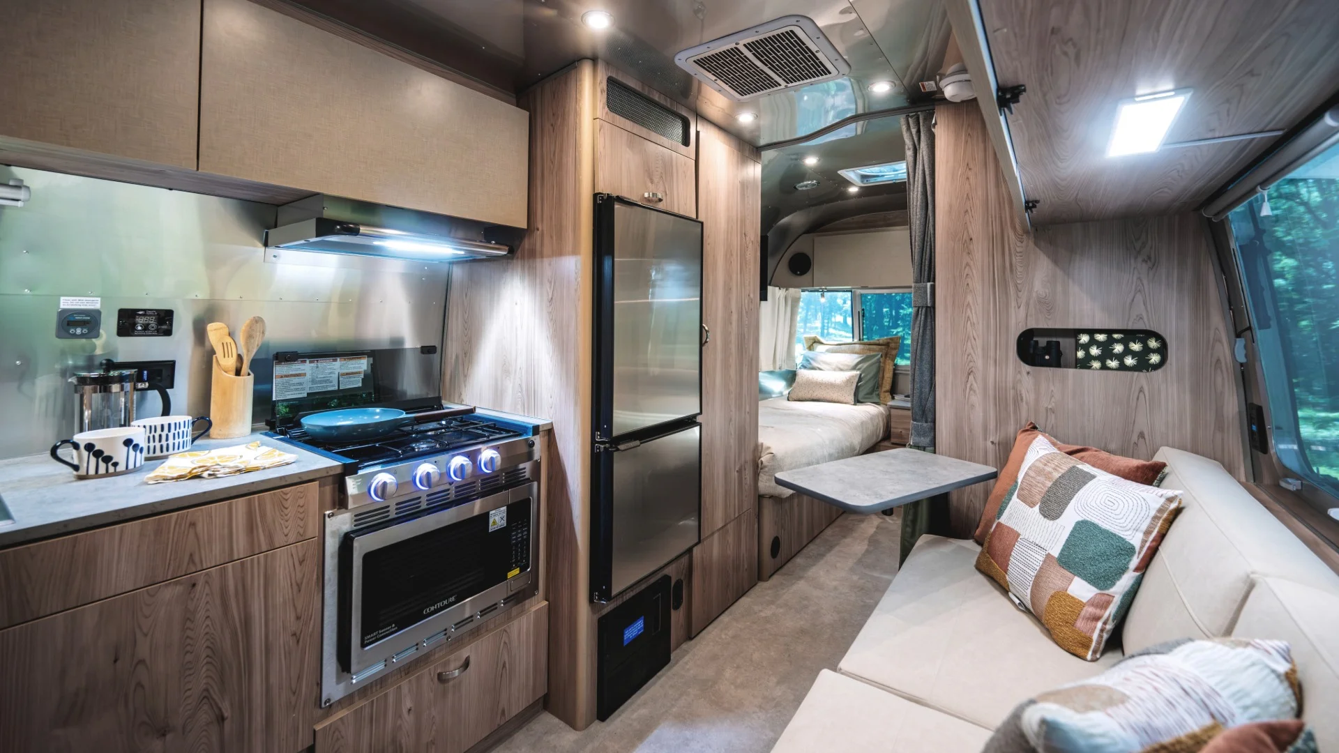 Modern RV interior with a compact kitchen, including a stove and fridge, and a cozy living area with a sofa, pillows, and a bed in the background.